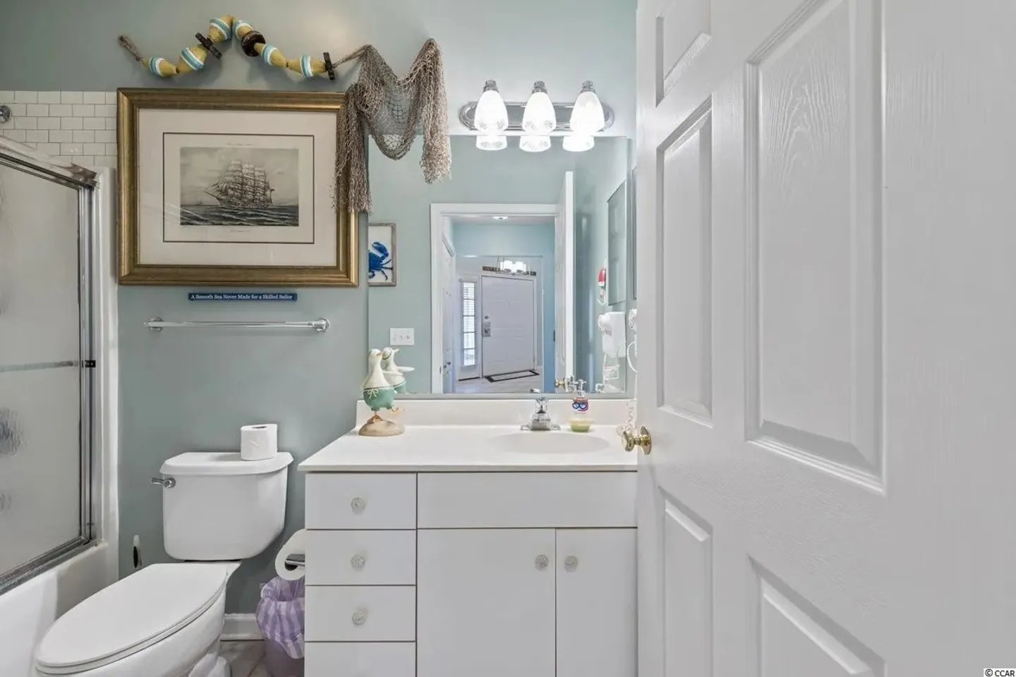 Bathroom with white vanity, toilet, mirror, framed ship art, and nautically themed wall decor.