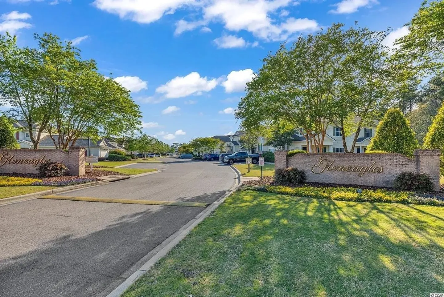 Neighborhood entrance with a paved road flanked by brick signs reading Gleneagles, surrounded by manicured lawns, trees, and residential buildings under a blue sky.