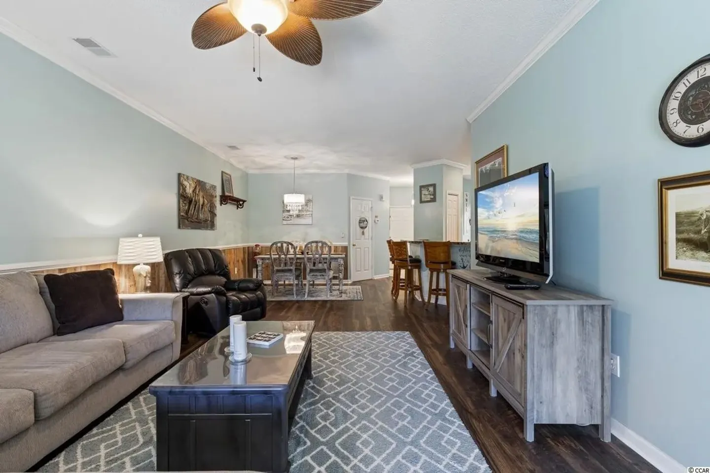 Open-concept living room with light blue walls, wood-look flooring, a sofa, coffee table, TV console, and a dining area in the background.