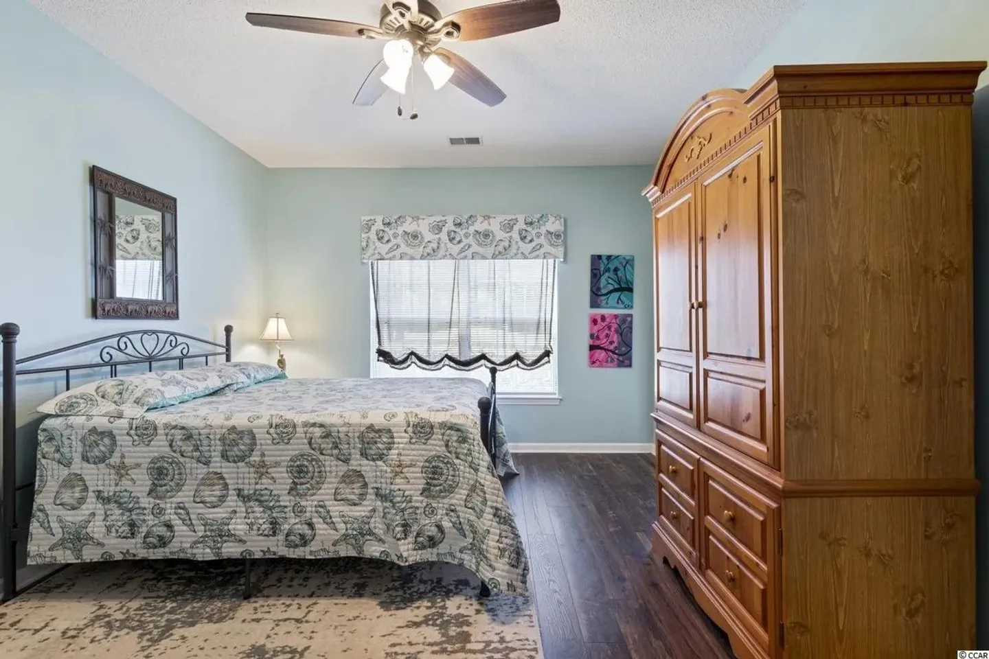 Bedroom with sea-life patterned bedspread, dark wood-look floors, a ceiling fan, and a large wooden armoire.