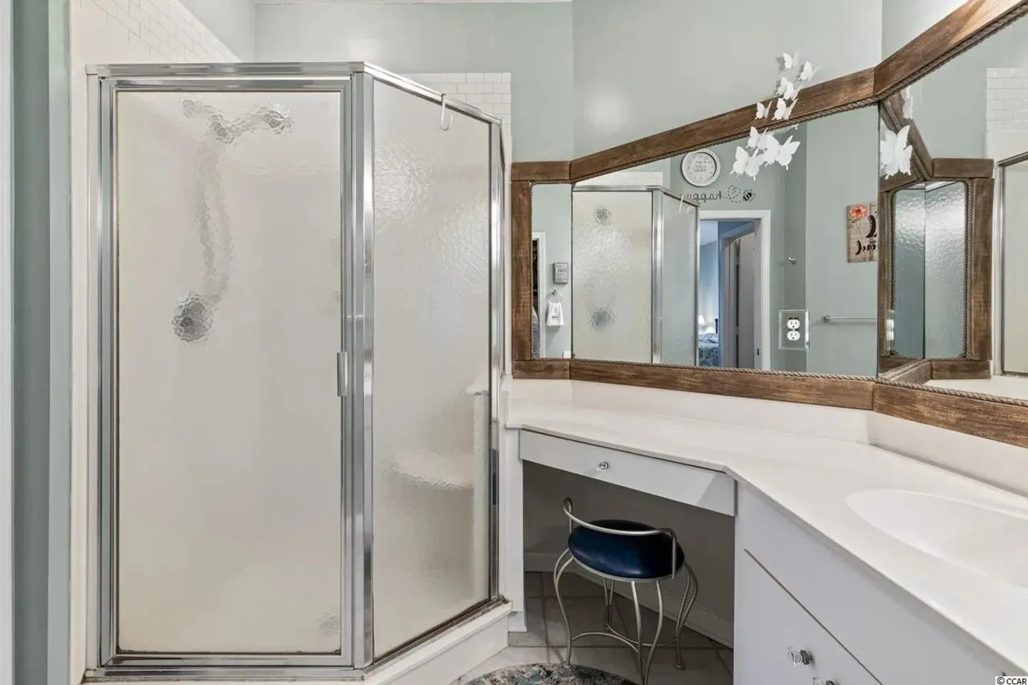 Bathroom showing a frosted glass shower stall next to a vanity with a sitting area, mirror, and stool.