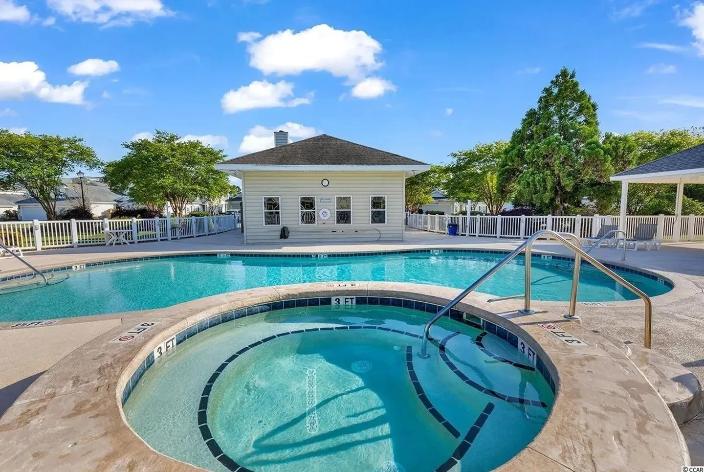 Outdoor swimming pool and hot tub area with a clubhouse building in the background under a blue sky.