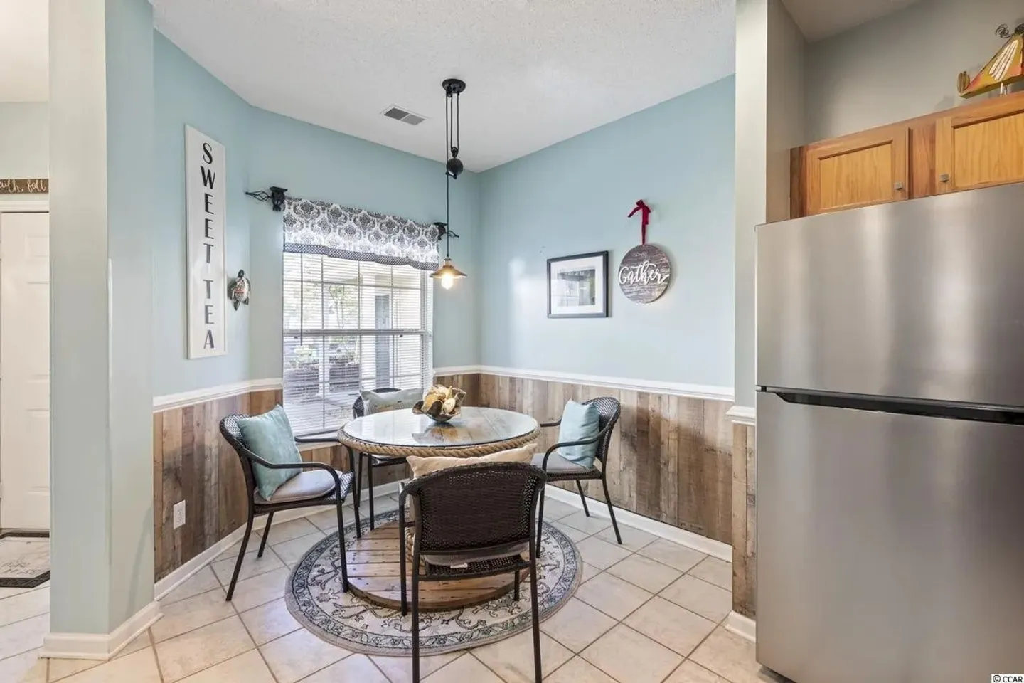 Dining nook with a round table, wicker armchairs, and light blue walls adjacent to a kitchen refrigerator.