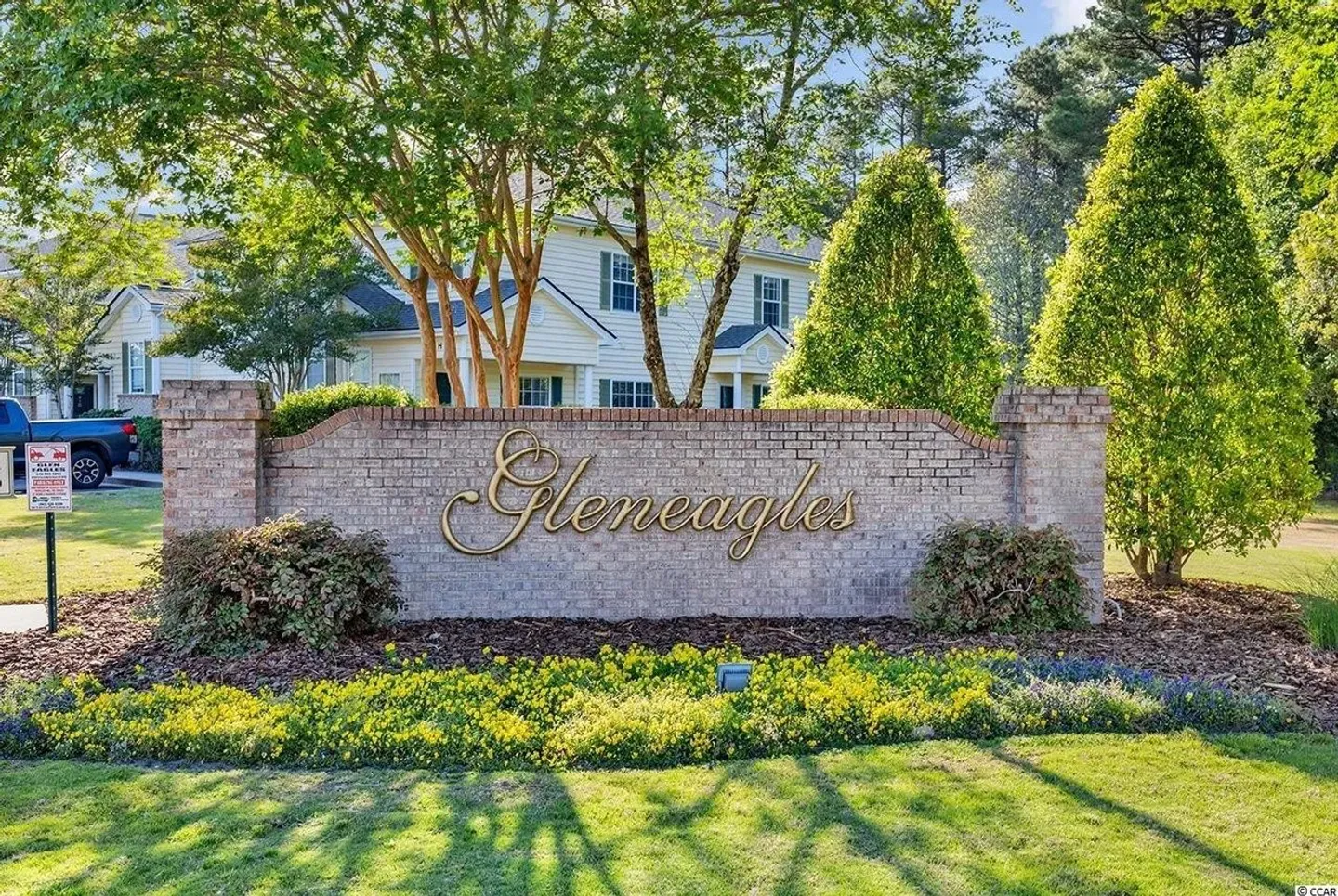 A curved brick entrance sign reading Gleneagles bordered by yellow flowers and green landscaping in front of residential buildings.