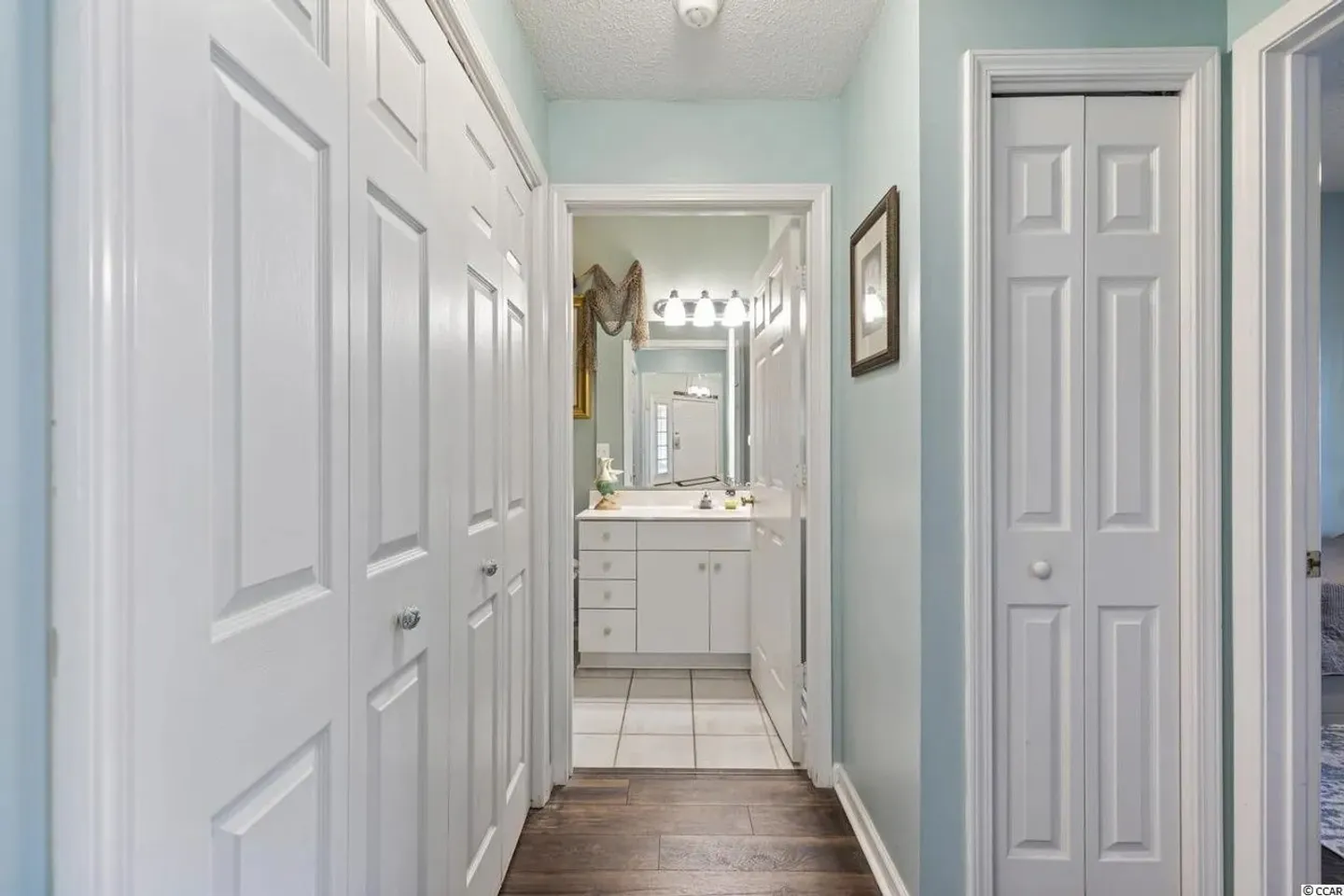 Hallway with light blue walls and white bifold closet doors leading to an ensuite bathroom with a white vanity.