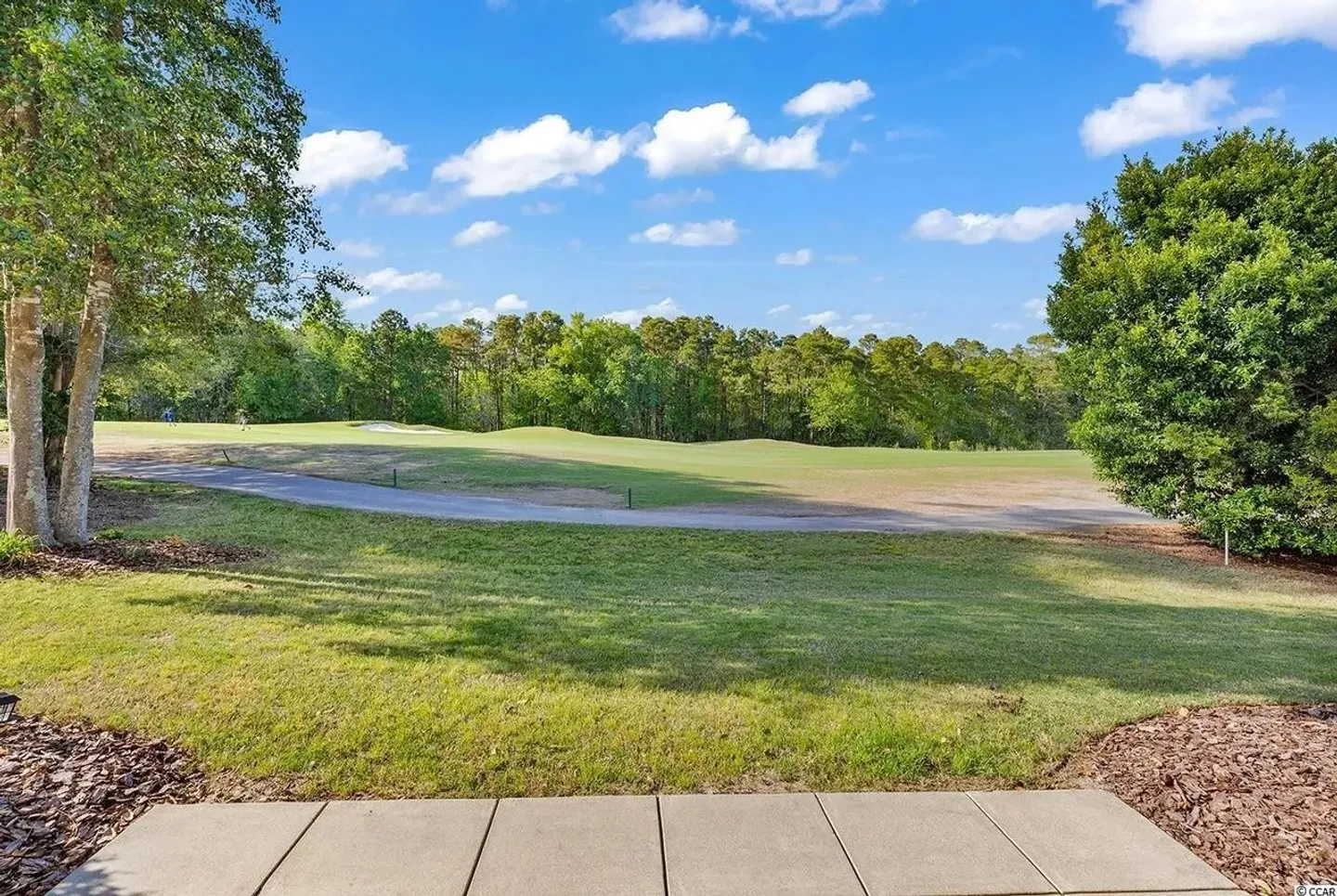 View from a paved patio overlooking a grassy lawn, a paved cart path, and a golf course fairway lined with trees under a blue sky.