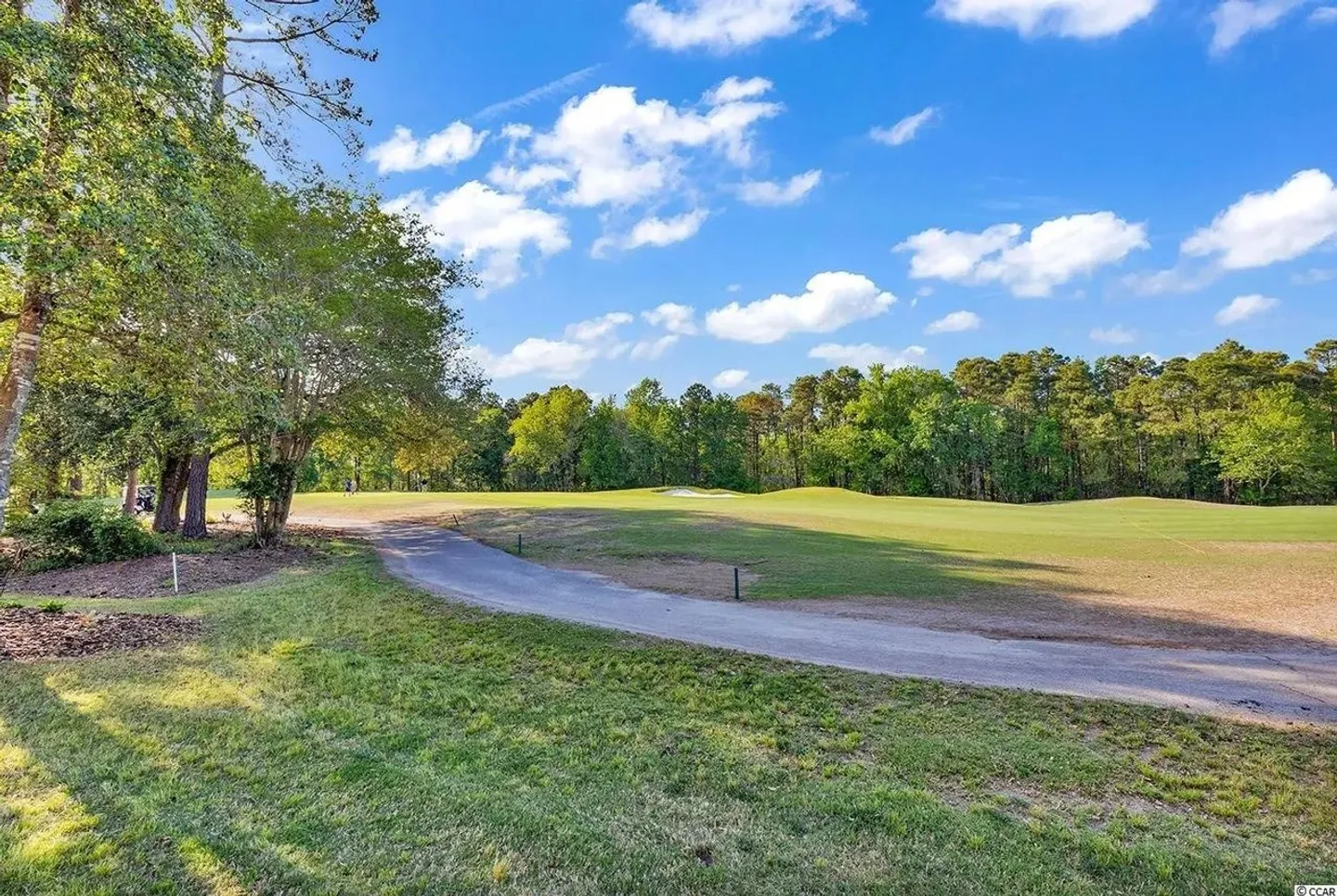 A paved cart path curving past green fairways bordered by mature trees under a blue sky with light clouds.