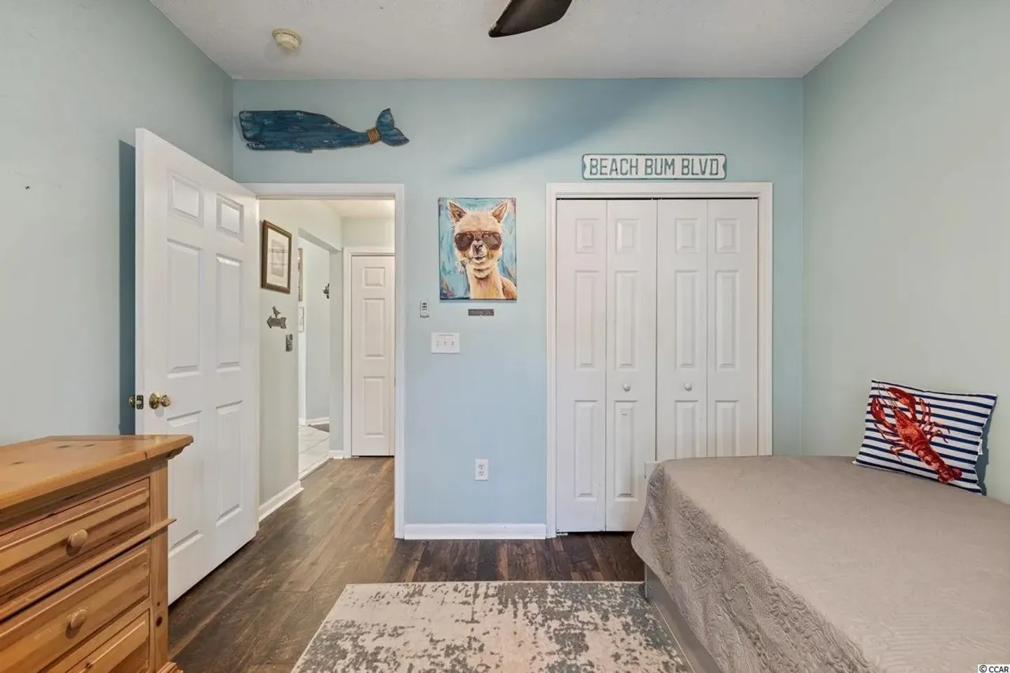 Bedroom with light blue walls, wooden dresser, closet with white bi-fold doors, and a bed with a lobster pillow.