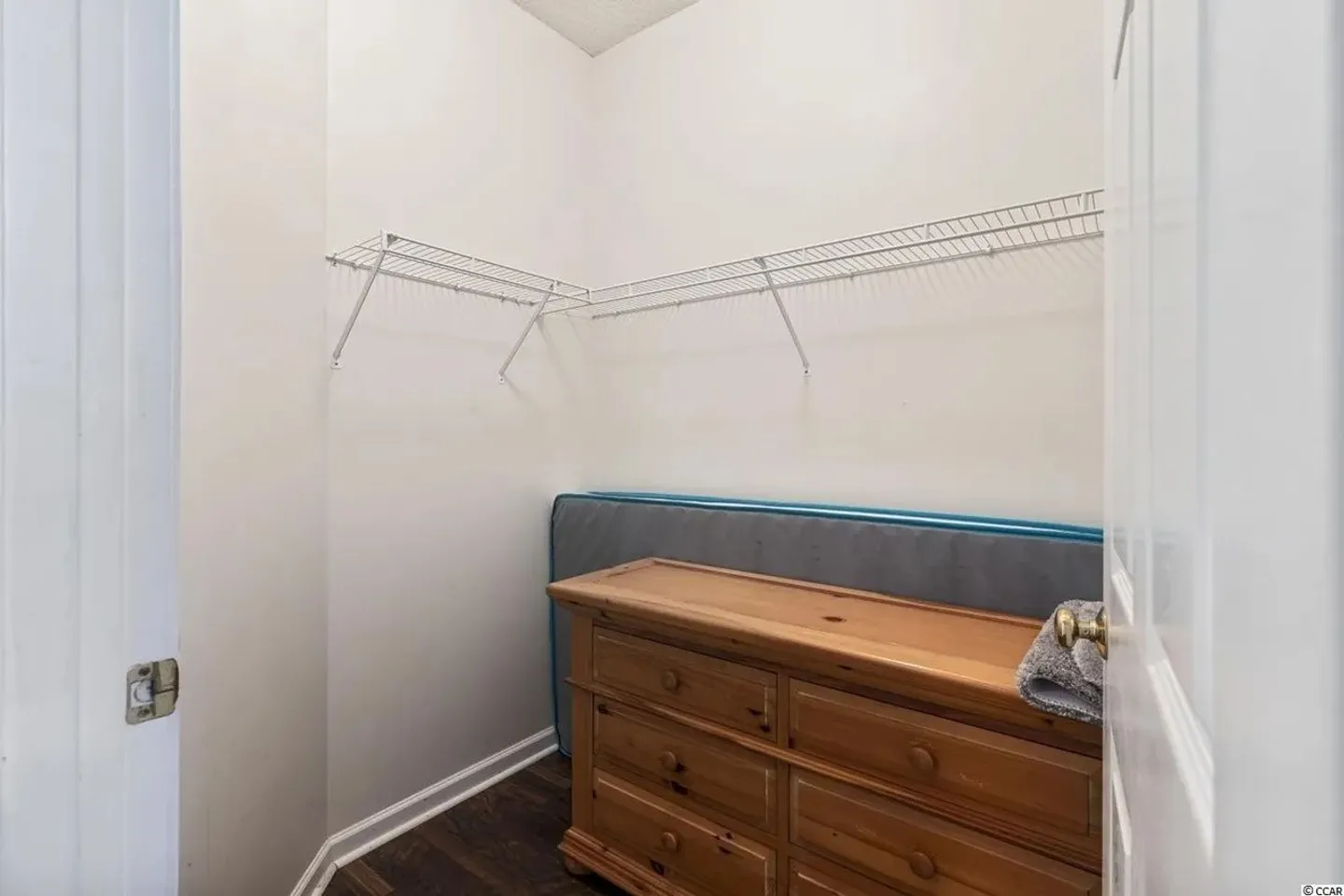A walk-in closet with dark wood-look flooring, white wire shelving, and a wooden chest of drawers.