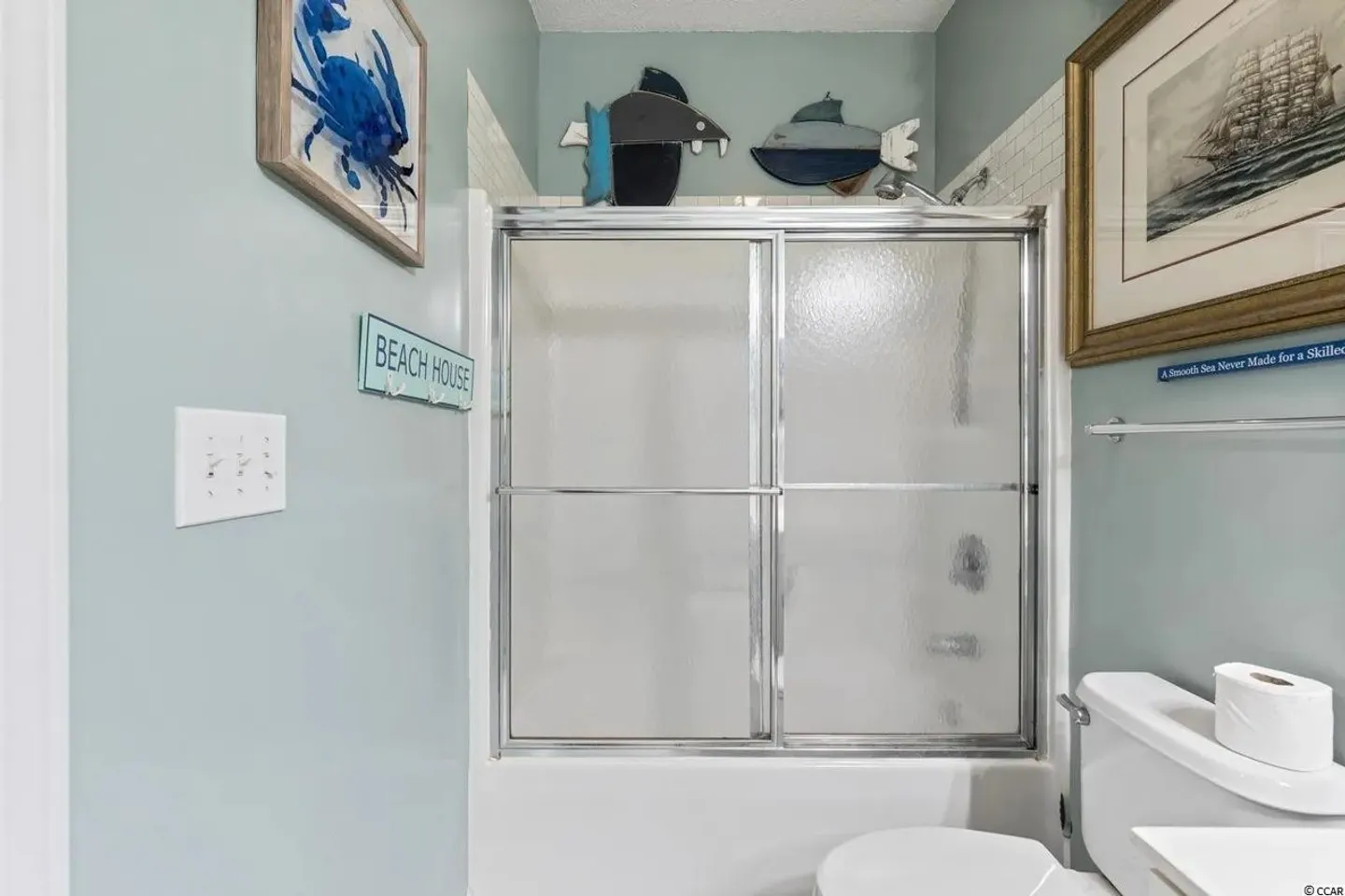 Bathroom showing a bathtub with frosted glass sliding doors, toilet, light blue walls, and coastal decor.