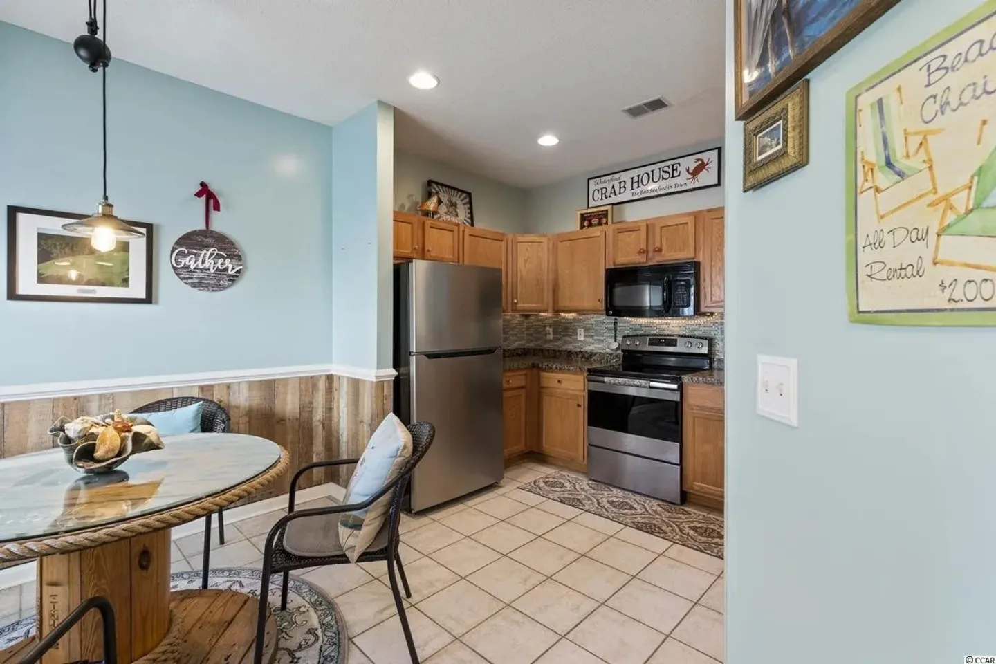 Dining area with a round glass-topped table beside an open kitchen with wooden cabinets and stainless steel appliances.