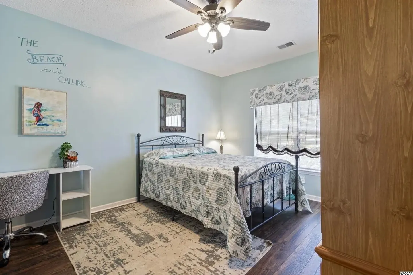 Bedroom with light blue walls, a metal frame bed with seashell-patterned bedding, a small white desk, and dark wood-style flooring.
