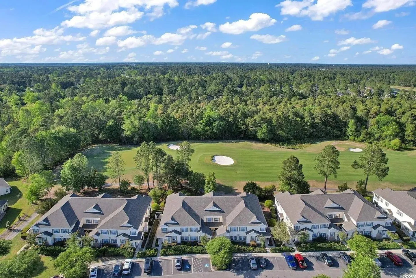 Aerial view of multi-story condominium buildings backing up to a green golf course surrounded by dense pine forest under a blue sky.