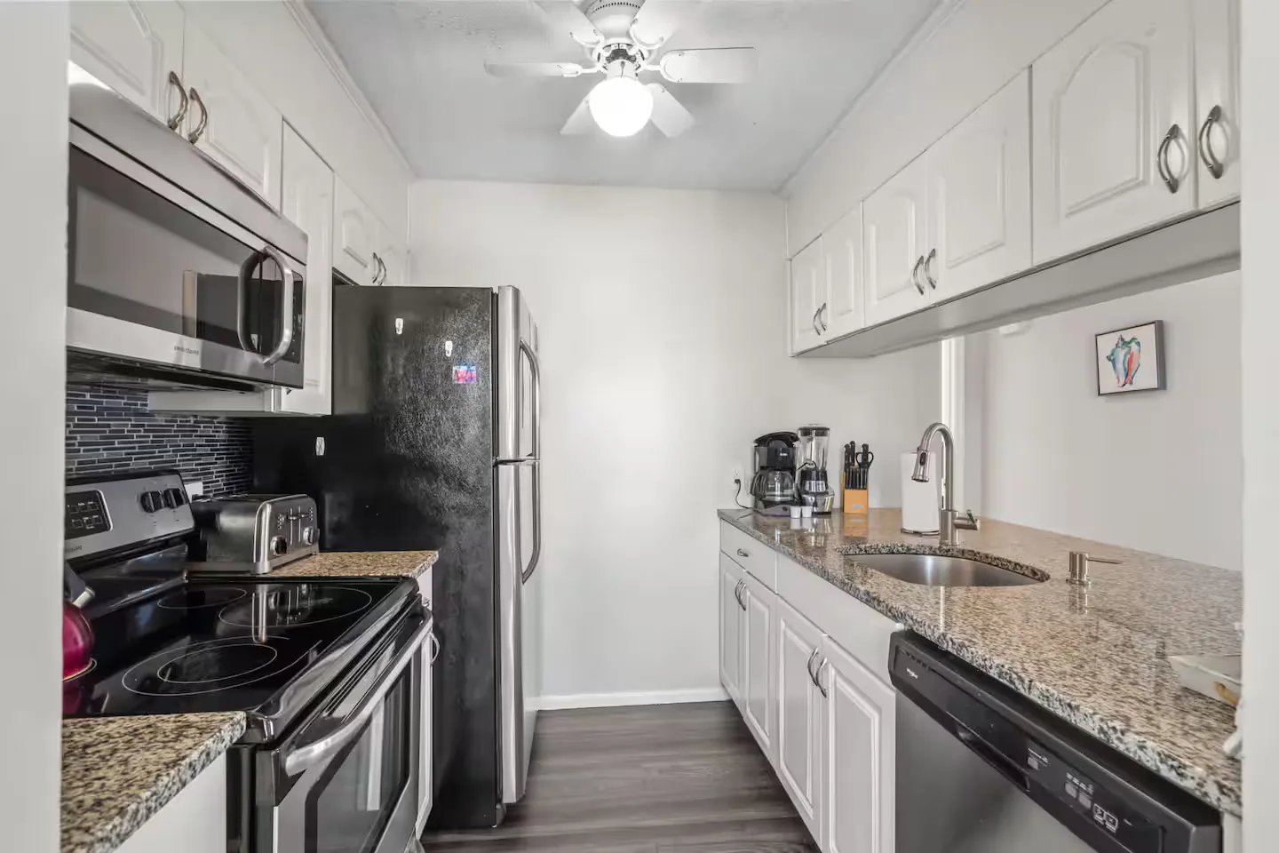 Galley-style kitchen with white cabinetry, granite-style countertops, stainless steel appliances, and dark wood-look flooring.