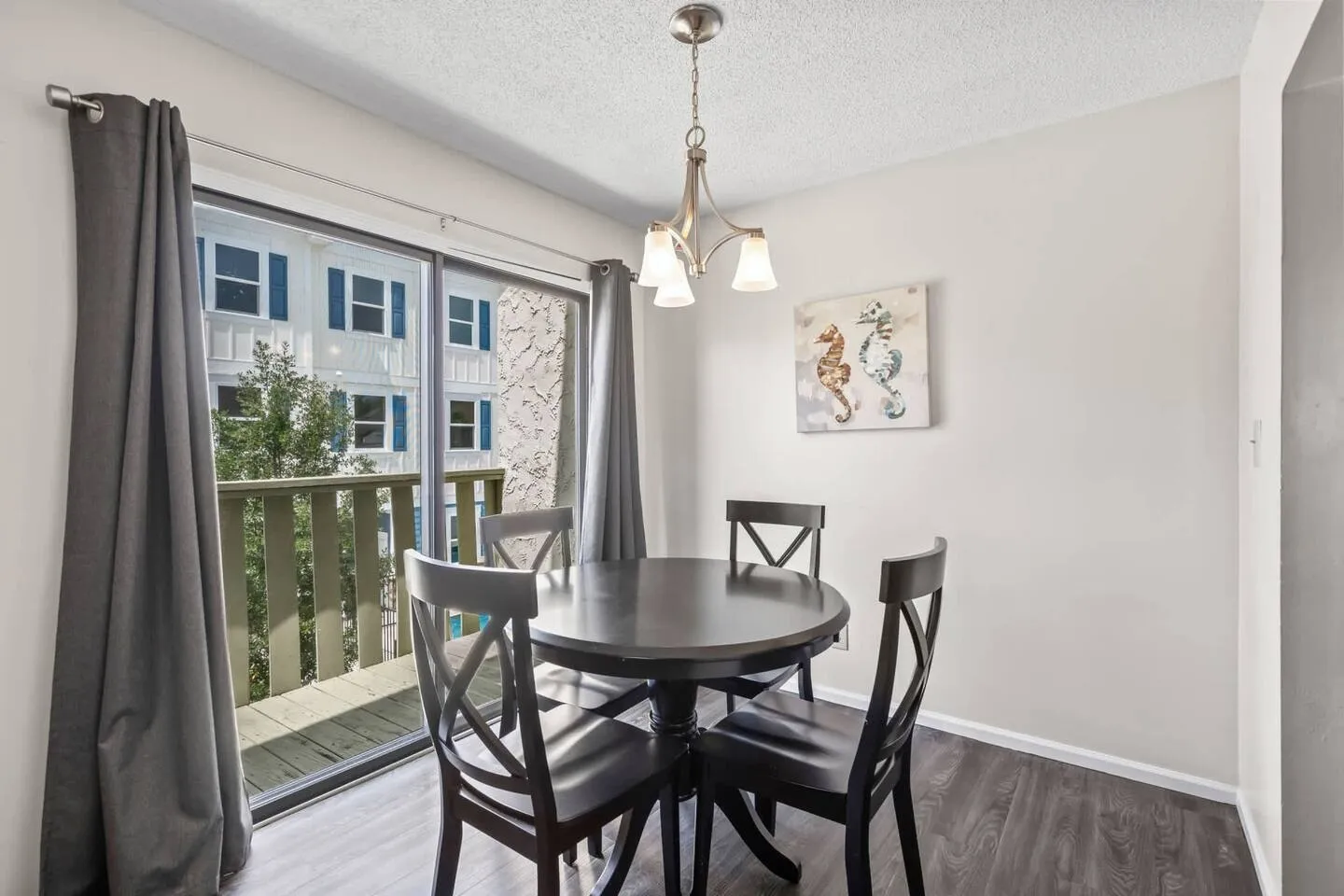 Dining area with a round dark wood table, four chairs, a hanging chandelier, and a sliding glass door leading to a balcony.