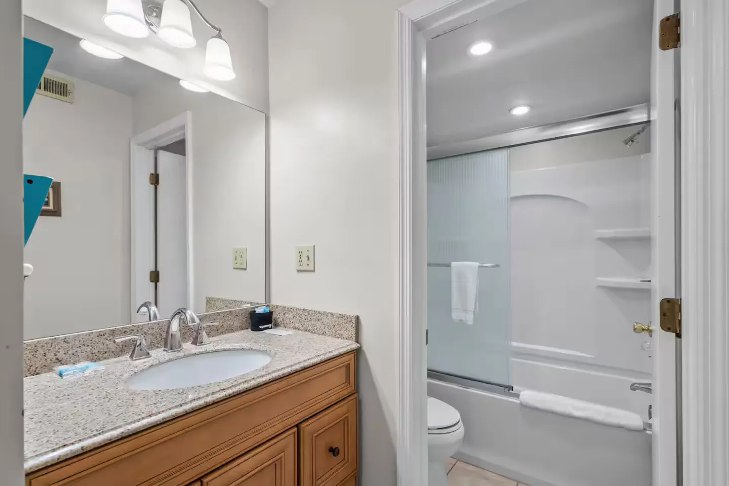 Bathroom vanity with speckled countertop and mirror leading into an adjoining room with a toilet and bathtub shower combo.