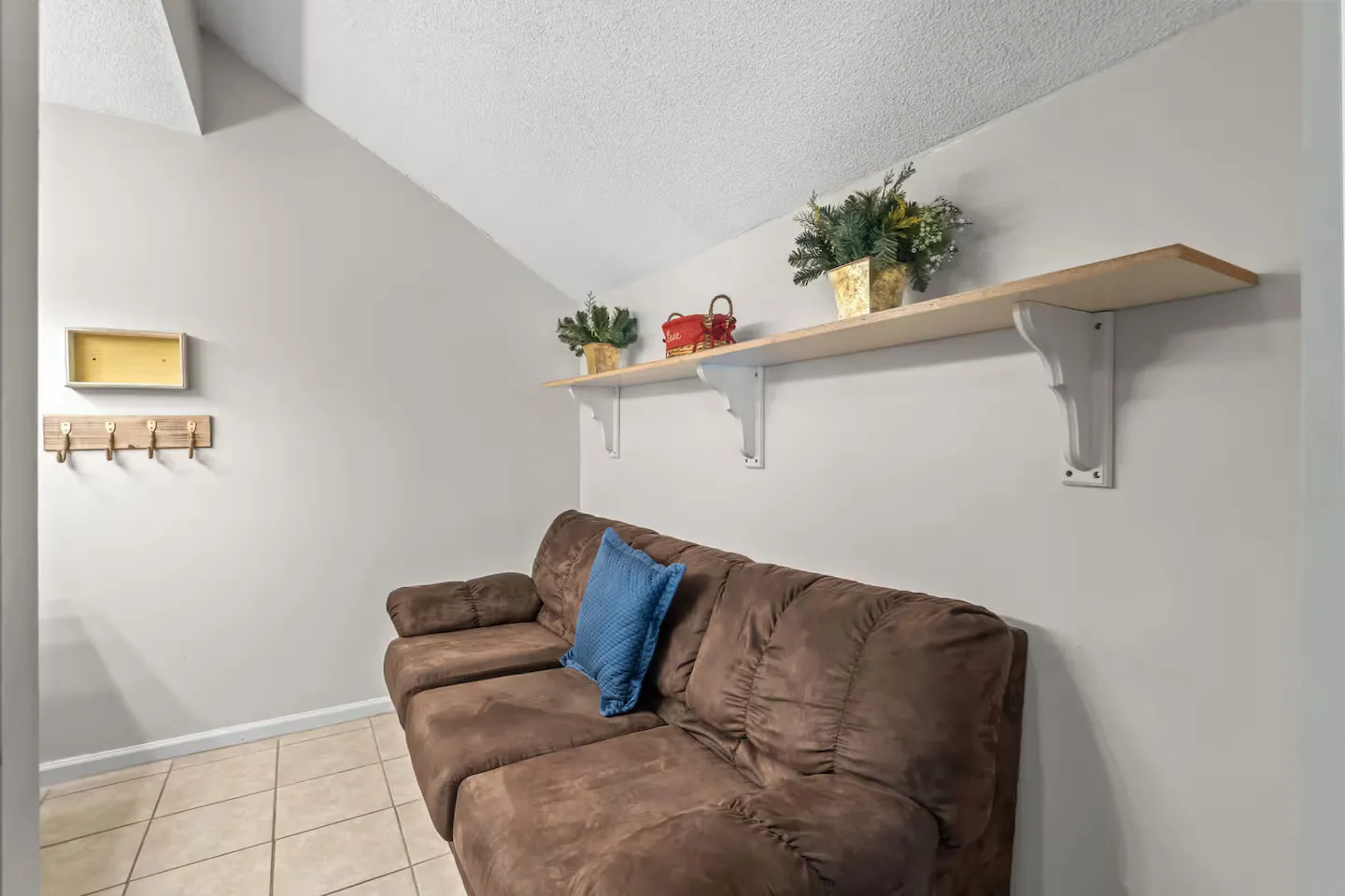 Brown sofa in a room with vaulted ceiling, tiled floor, a wall shelf with decor, and coat hooks.