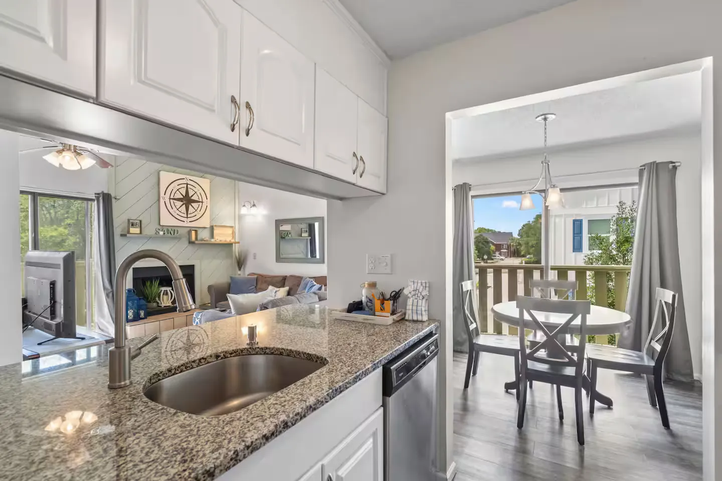 Kitchen counter and sink looking through a pass-through into the living room and an adjacent dining area with a table and chairs by a balcony.
