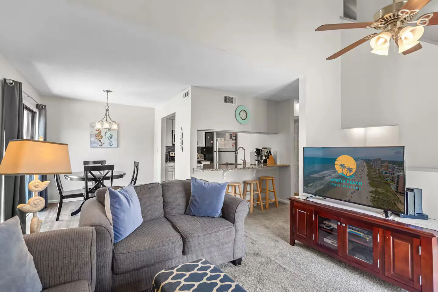Open living area with grey upholstered seating, a television on a wooden console, and a view into the dining area and kitchen.