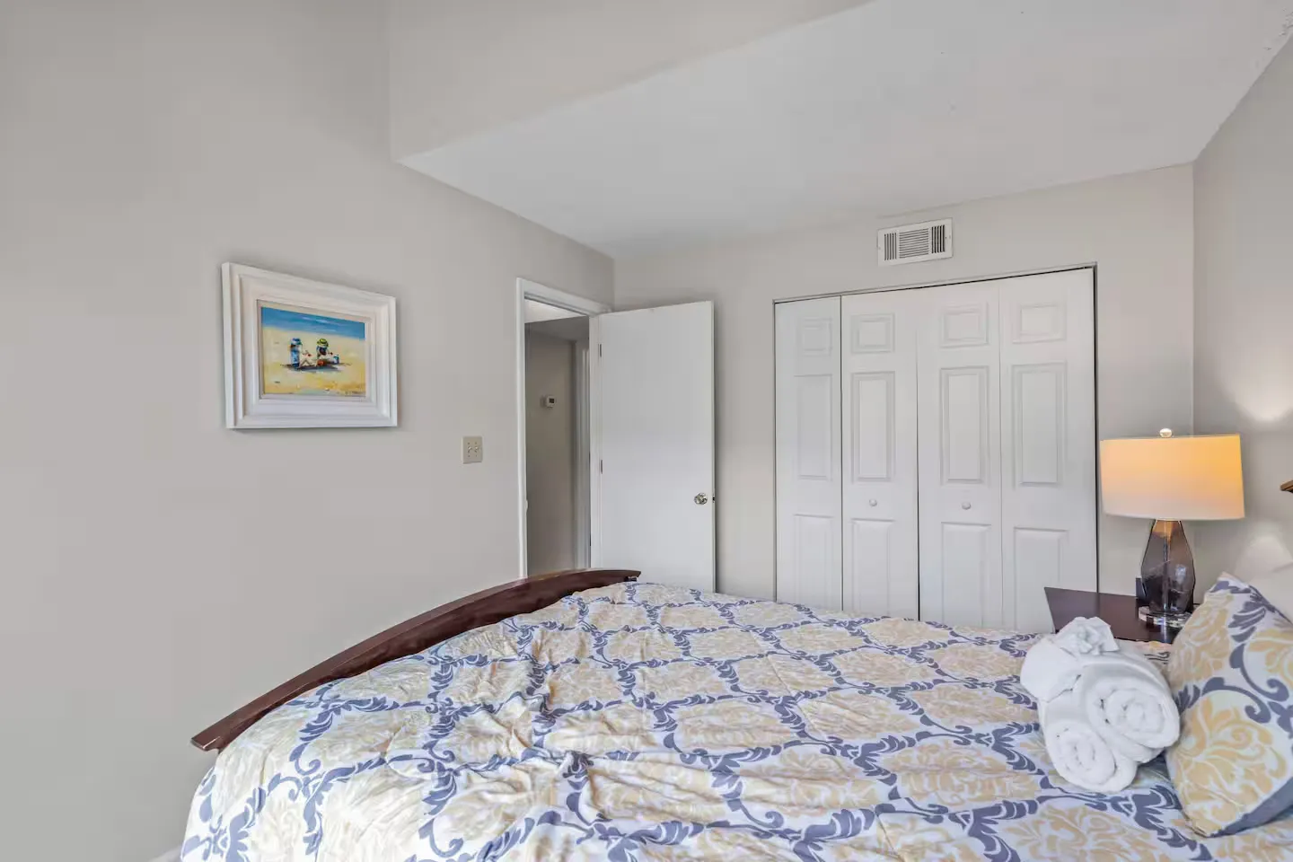 Bedroom featuring a patterned bedspread, rolled white towels, a framed picture on the wall, a bedside lamp, and white closet doors.