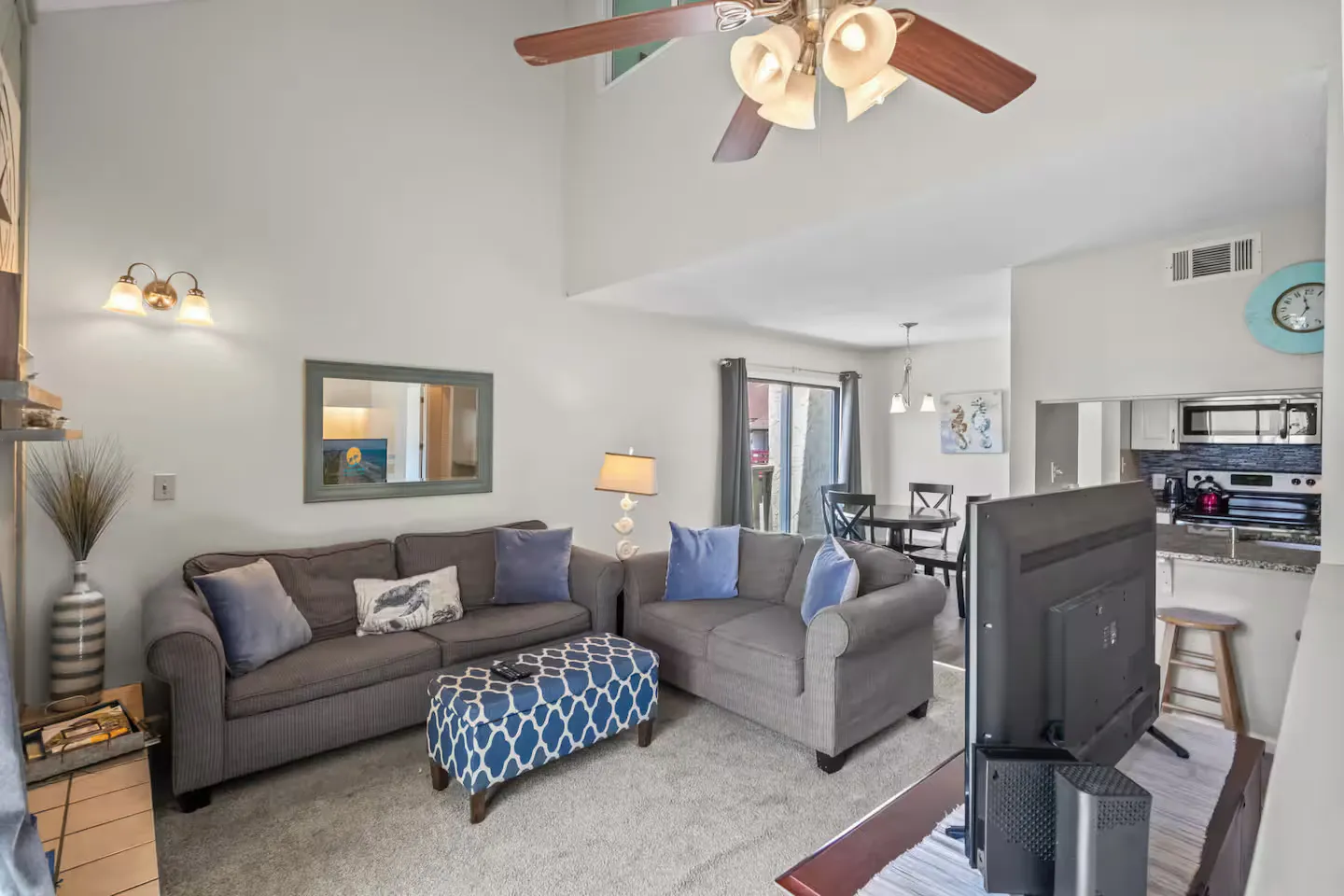 Living room with gray sofas, a patterned blue ottoman, a television, and an adjacent dining area.