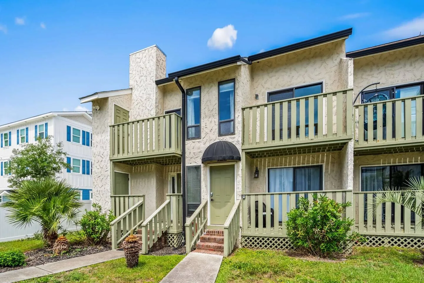 Exterior front view of a two-story beige townhome with pale green railings, balconies, and a front entry door with a small black awning.