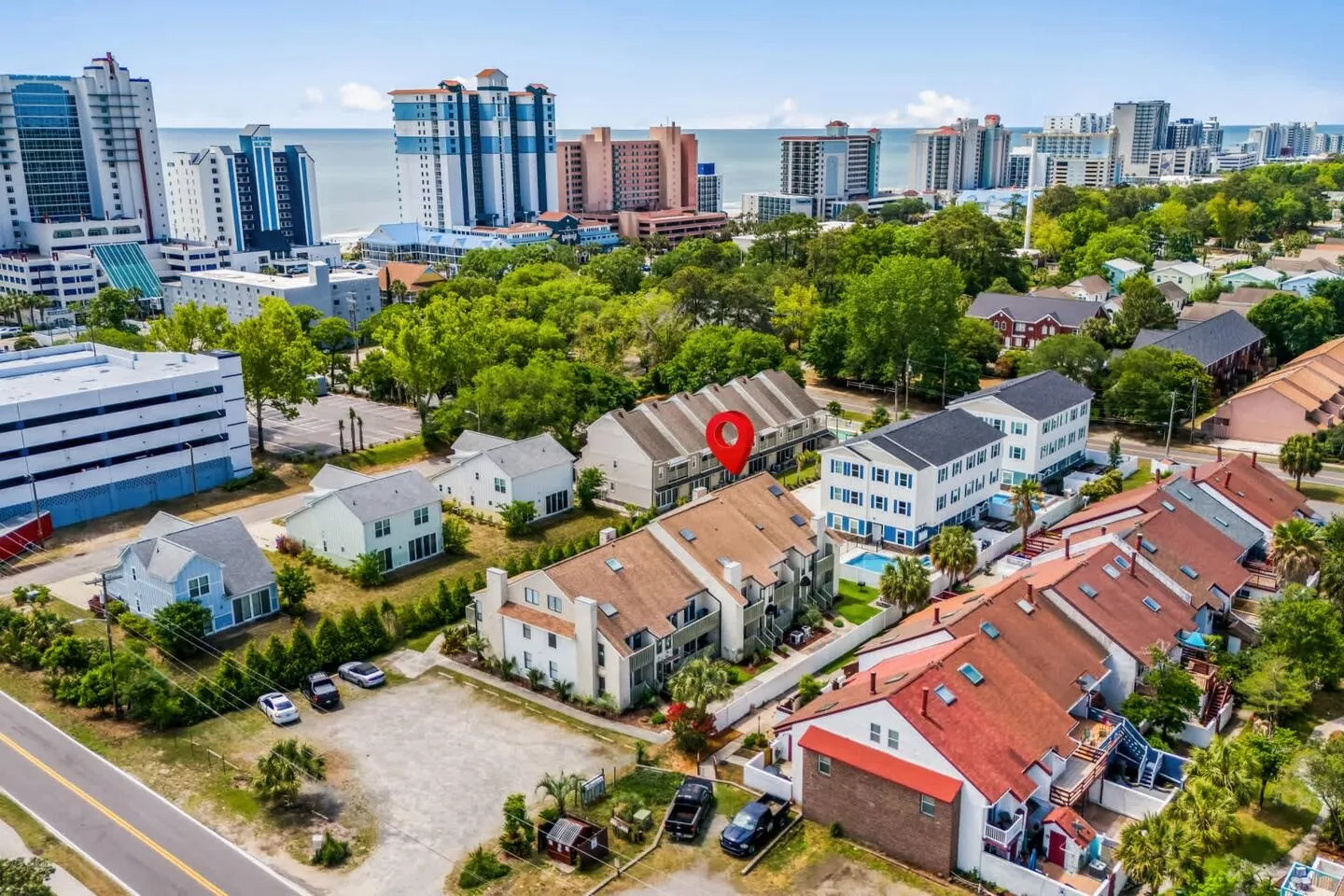 Aerial view of a coastal neighborhood with townhomes and high-rise buildings near the ocean, with a red location pin highlighting one complex.