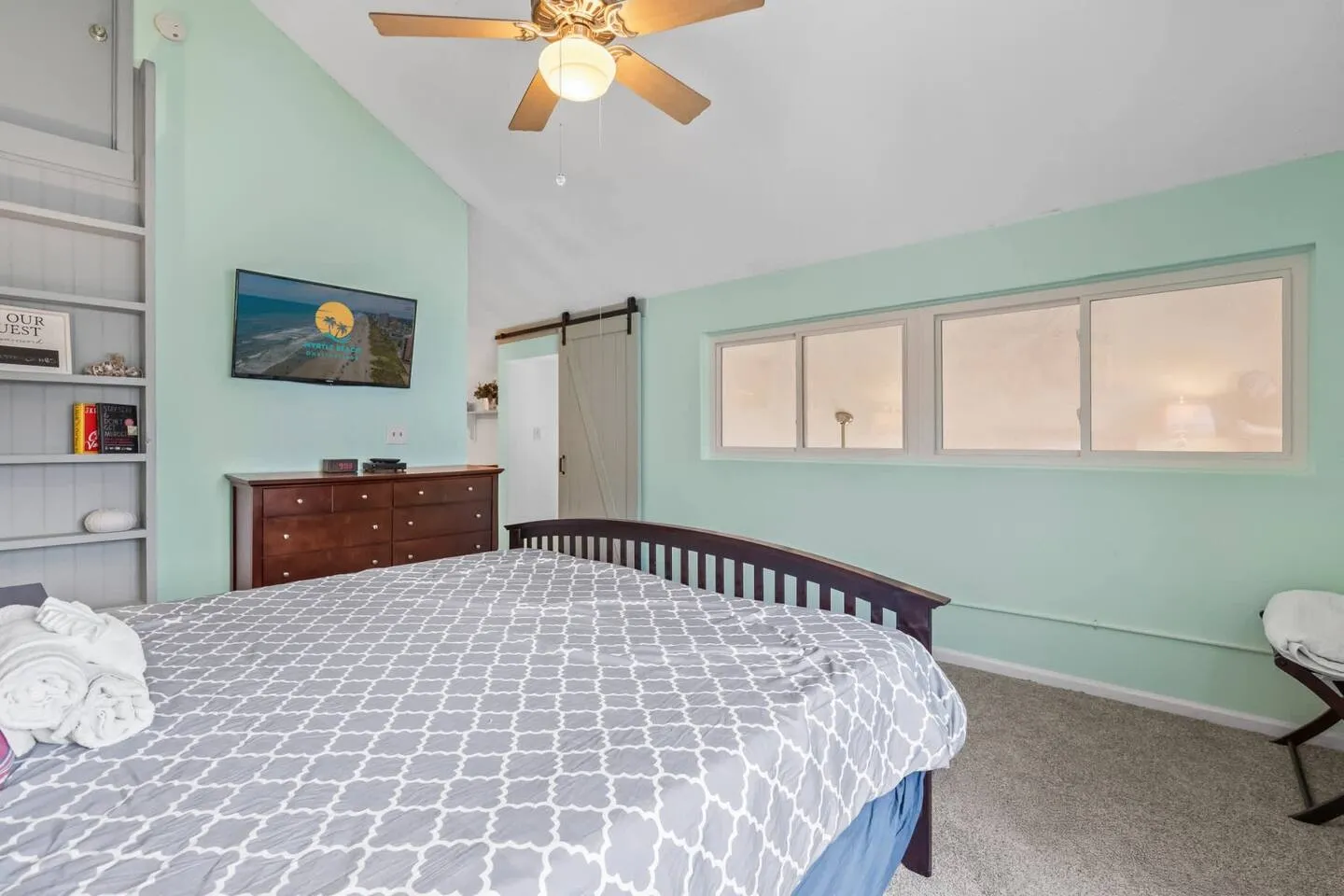 Bedroom with vaulted ceiling, light teal walls, a dark wood bed with patterned bedding, a ceiling fan, and frosted transom windows.