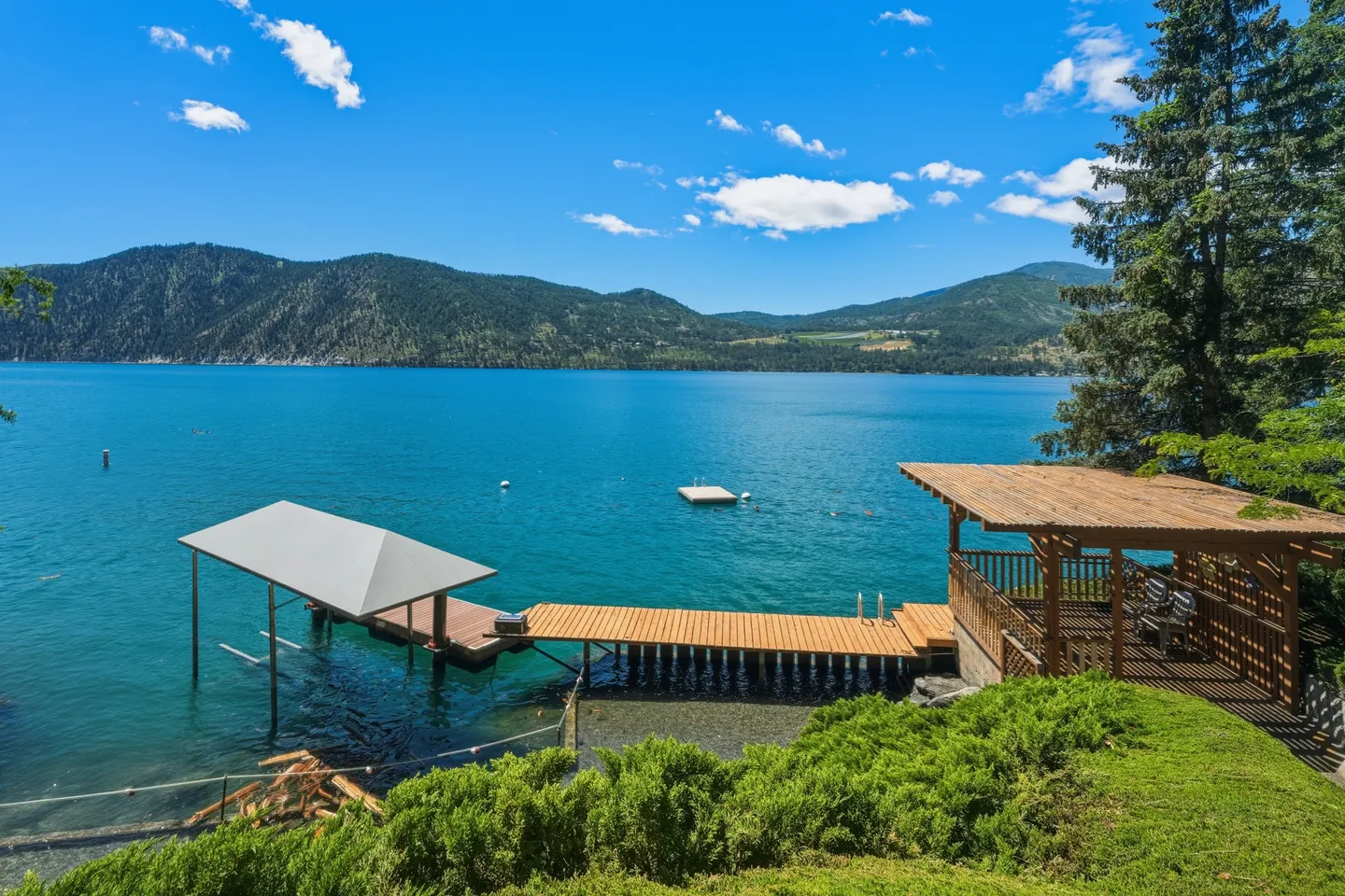 Elevated view of a lake dock, covered boat slip, and open-air gazebo overlooking blue water and forested mountains.