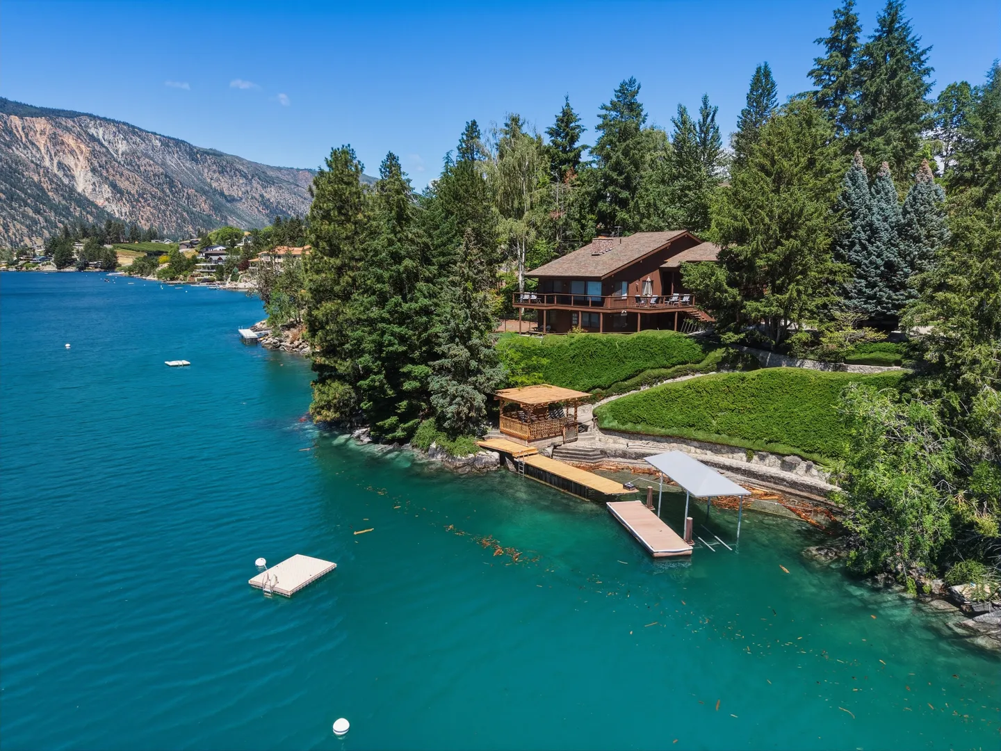 Aerial view of a lakefront house surrounded by pine trees with private docks, a covered boat slip, and turquoise water.