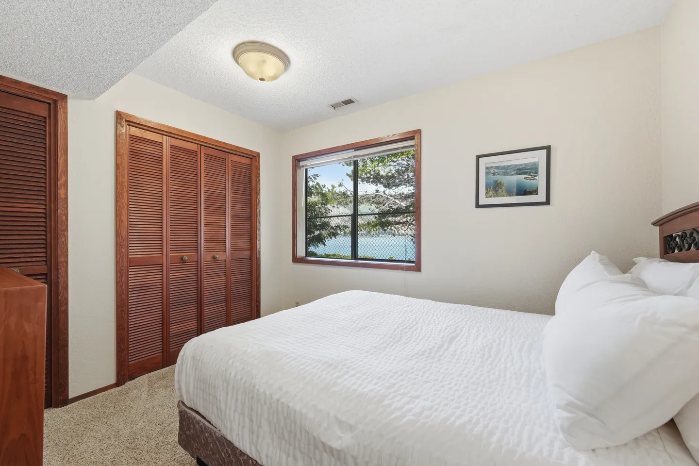 Bedroom with a bed made with white linens, carpeted floor, wooden louvered closet doors, and a window with water views.