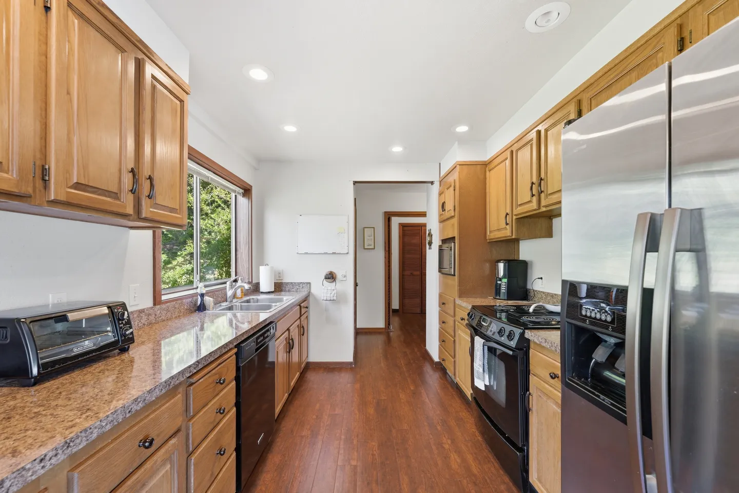 Galley-style kitchen featuring wood cabinetry, speckled countertops, stainless steel appliances, and wood-look flooring.