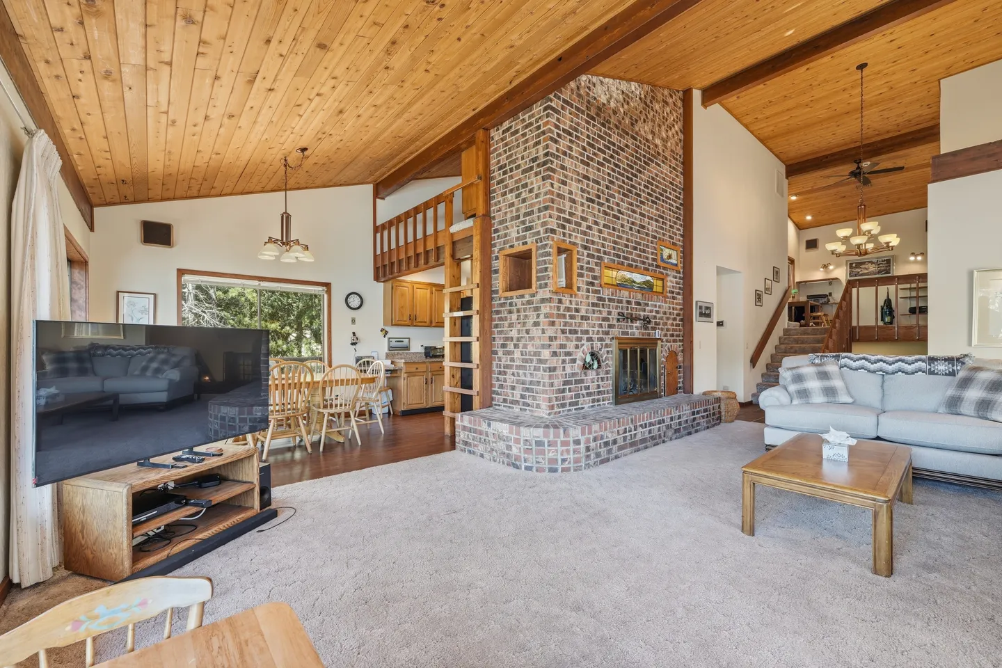 Open living area with wood-plank ceilings, a central brick fireplace, carpeted seating area, and dining space.