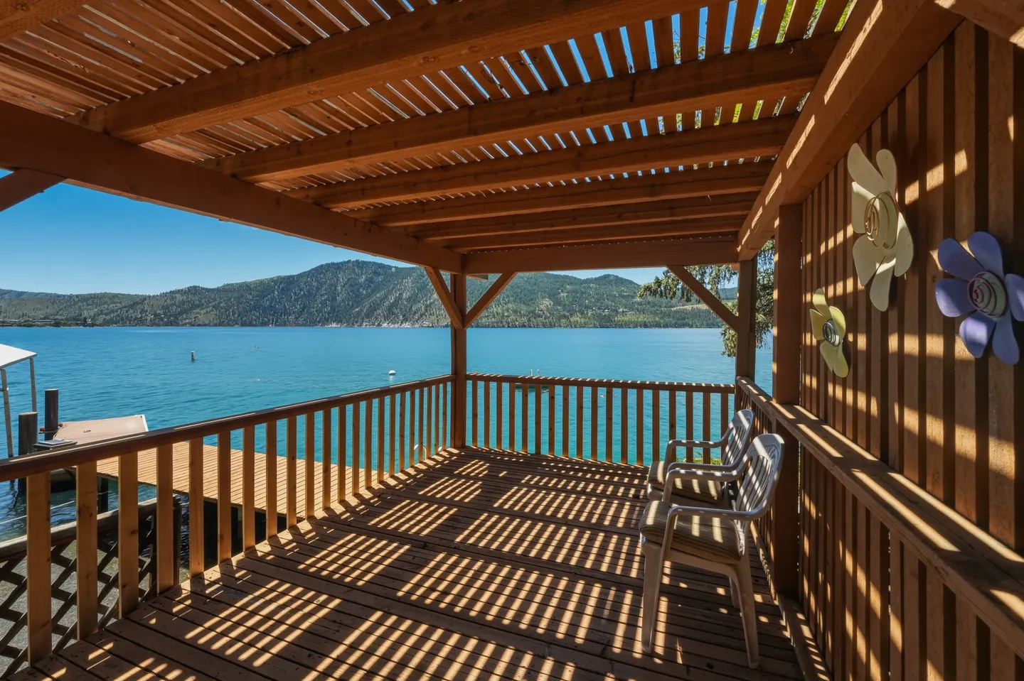 A covered wooden deck with a slatted pergola roof and seating, overlooking a wide blue lake and forested mountains under a clear sky.