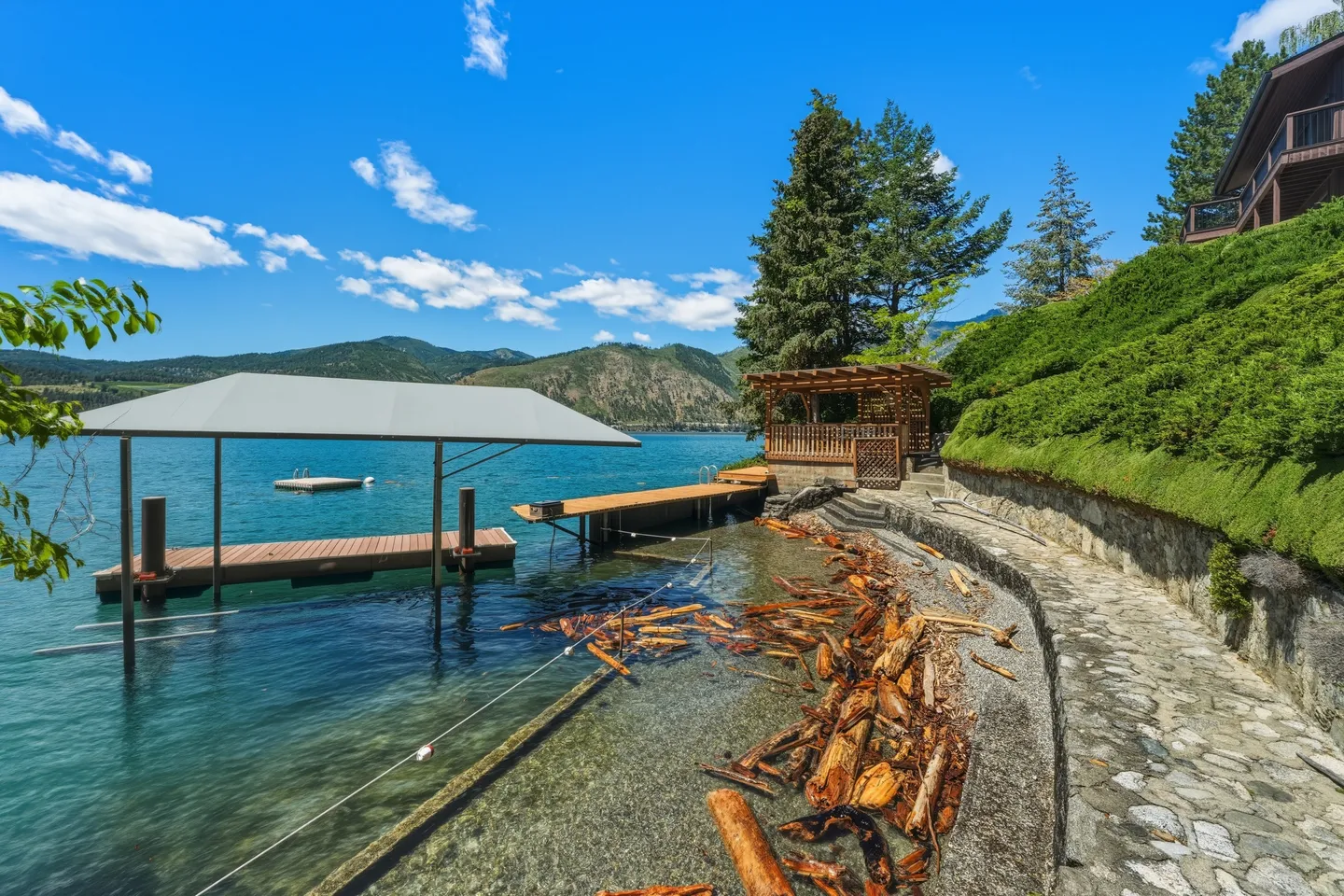 A private boat dock with a canopy shelter on a clear lake, beside a stone shoreline walkway and wooden gazebo.