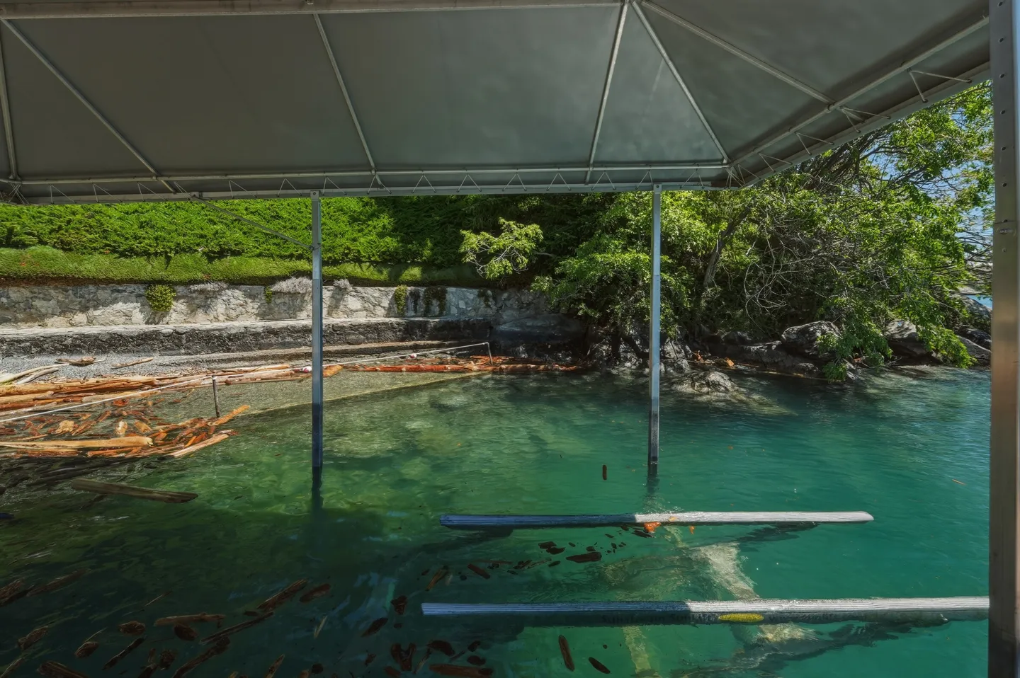 View from beneath a covered boat slip looking out onto clear water, a stone shoreline, and dense green vegetation.