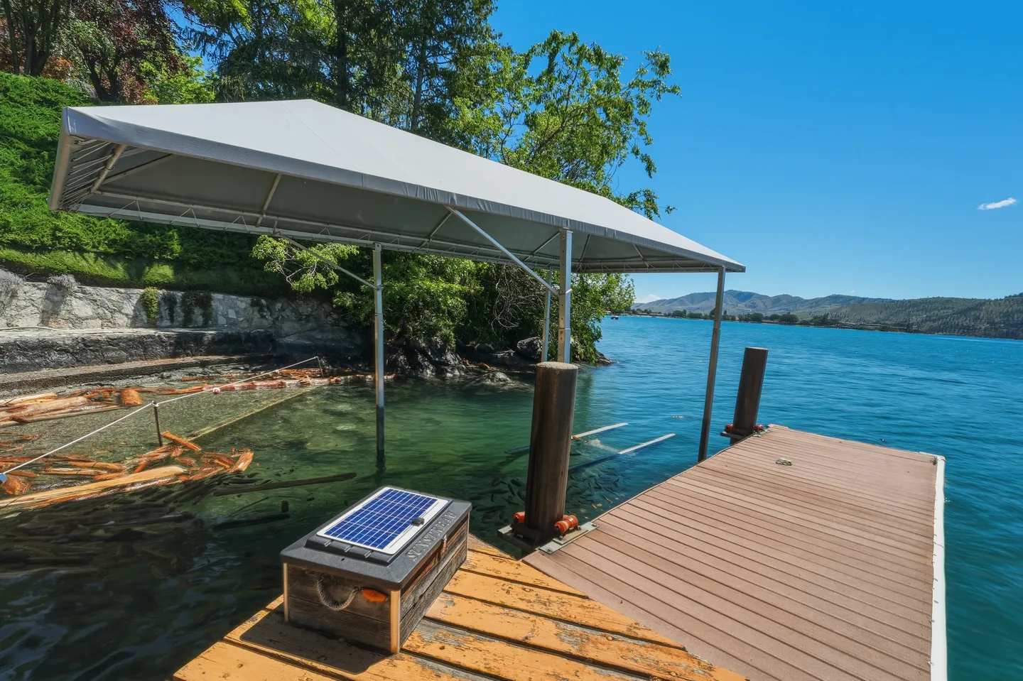 A wooden dock and covered boat lift sitting in clear blue water on a sunny day with distant hills.