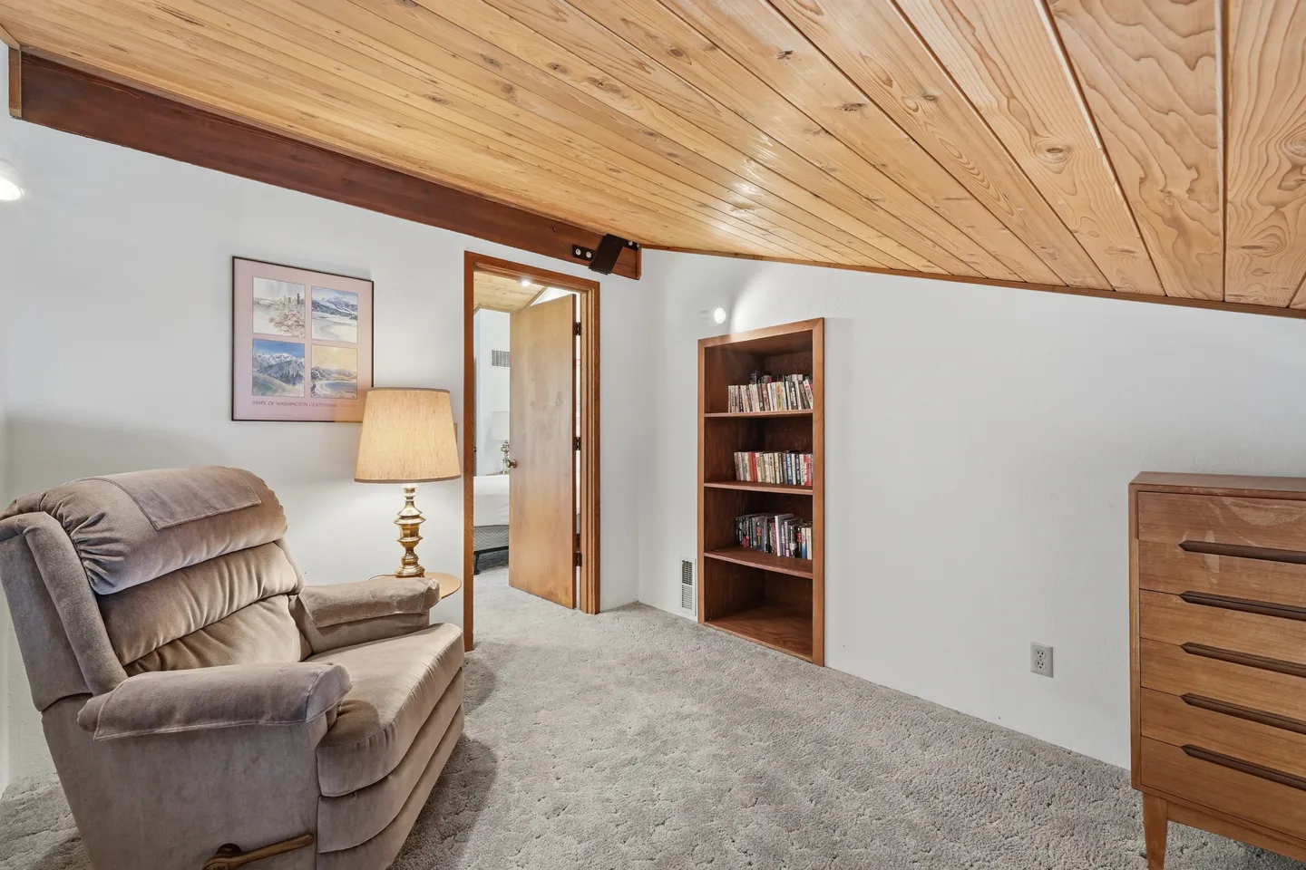 A carpeted room with a sloped wood-paneled ceiling, a plush recliner, a built-in bookcase, and a chest of drawers.