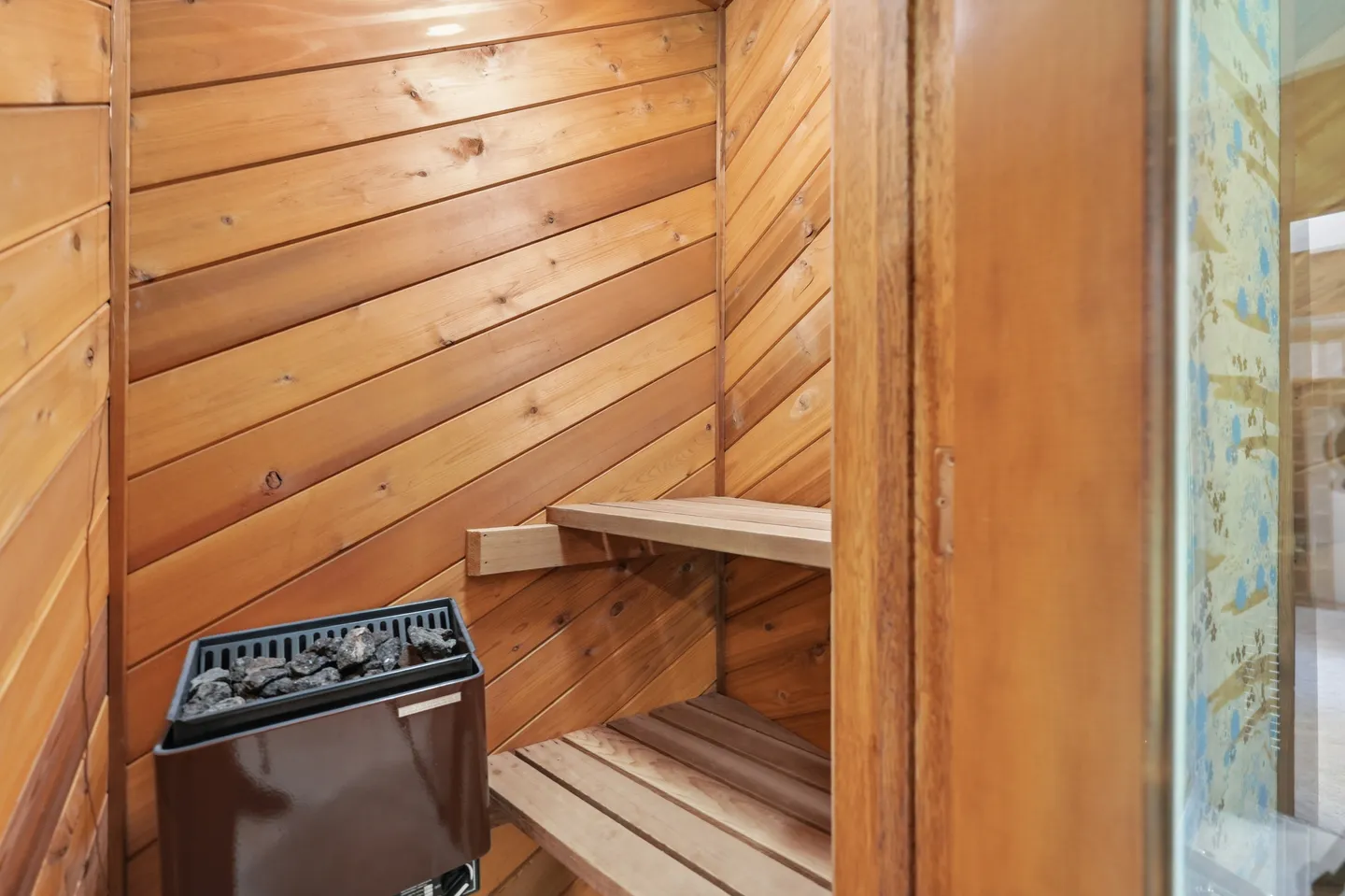An indoor wooden sauna with diagonal wood paneling, tiered benches, and an electric rock heater.