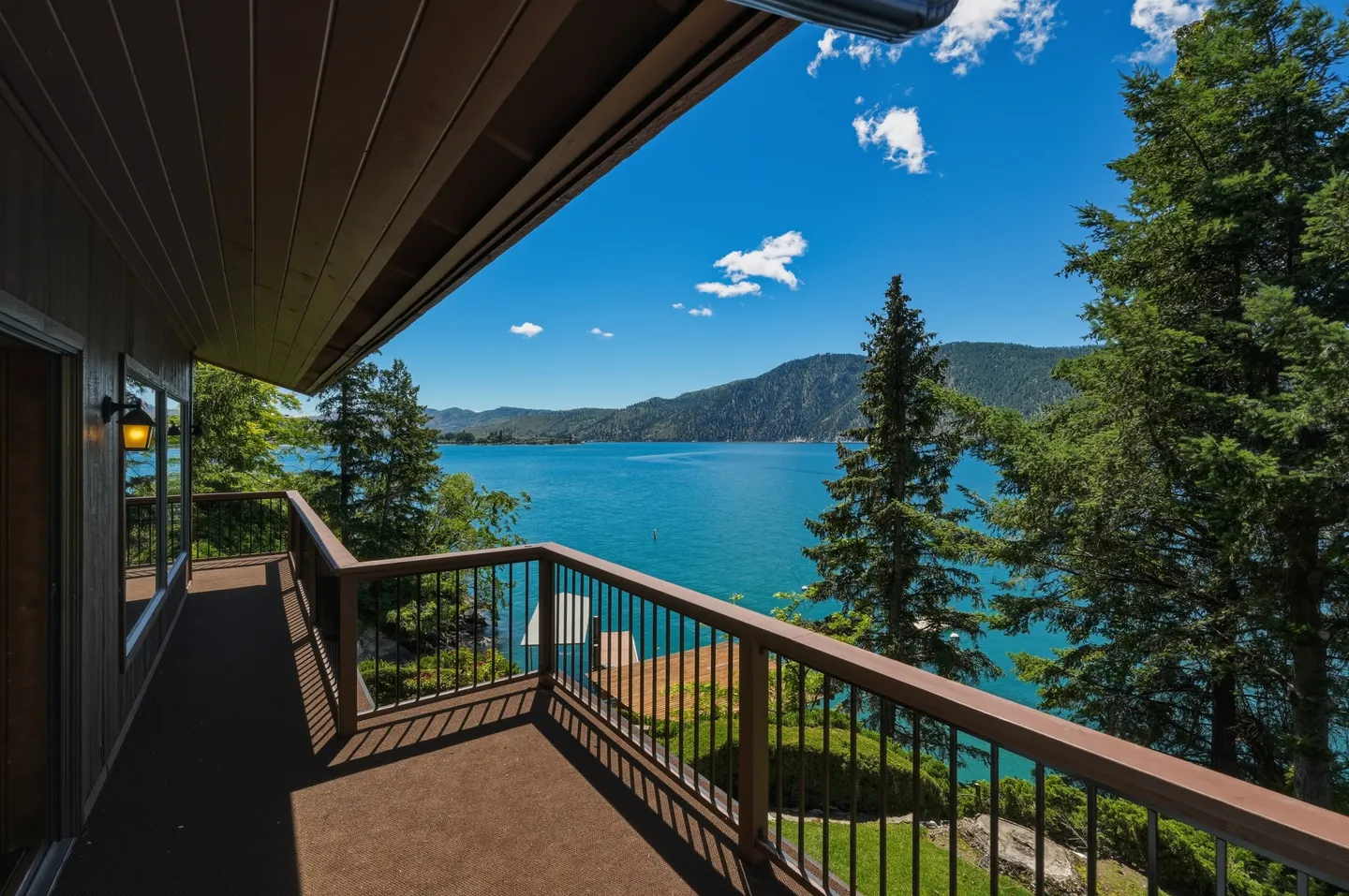 A covered balcony with dark brown railing overlooks a bright blue lake surrounded by mountains and pine trees.