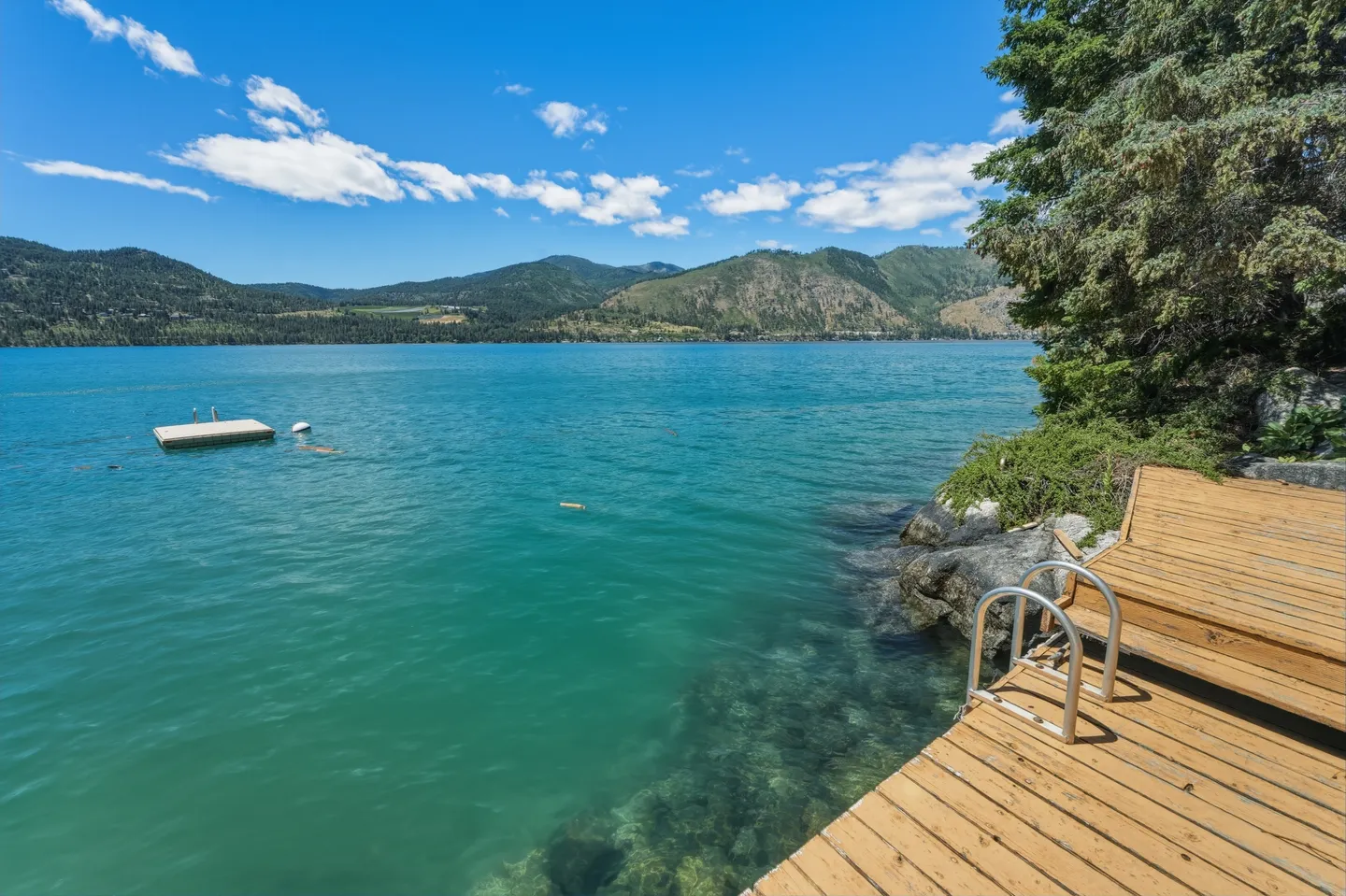 A wooden dock with a swim ladder sits on the edge of clear turquoise water overlooking distant mountains.