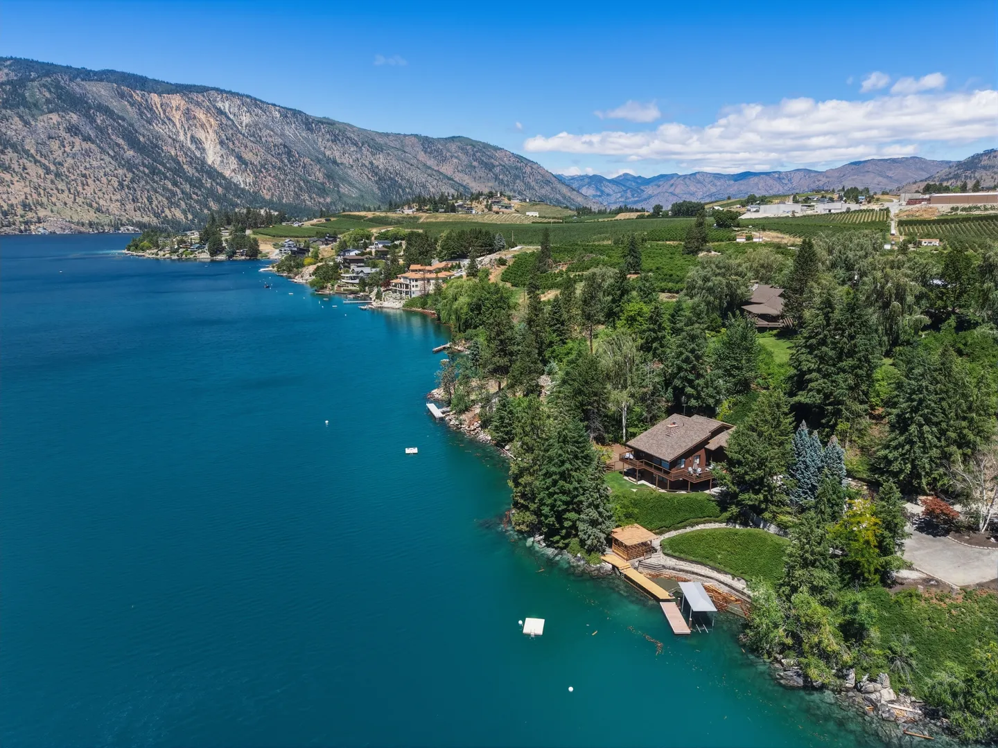 Aerial view of a lakefront house surrounded by green trees along deep blue water with mountains in the background.