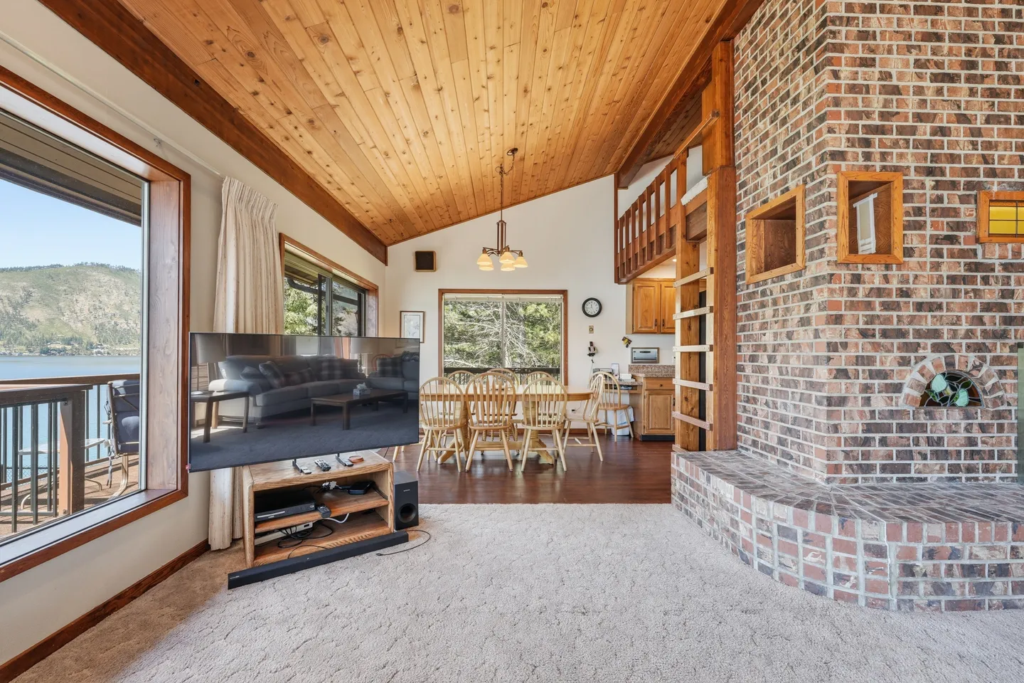 Open living area with wood-plank vaulted ceiling, brick fireplace structure, flat-screen TV, dining table, and windows showing a lake view.