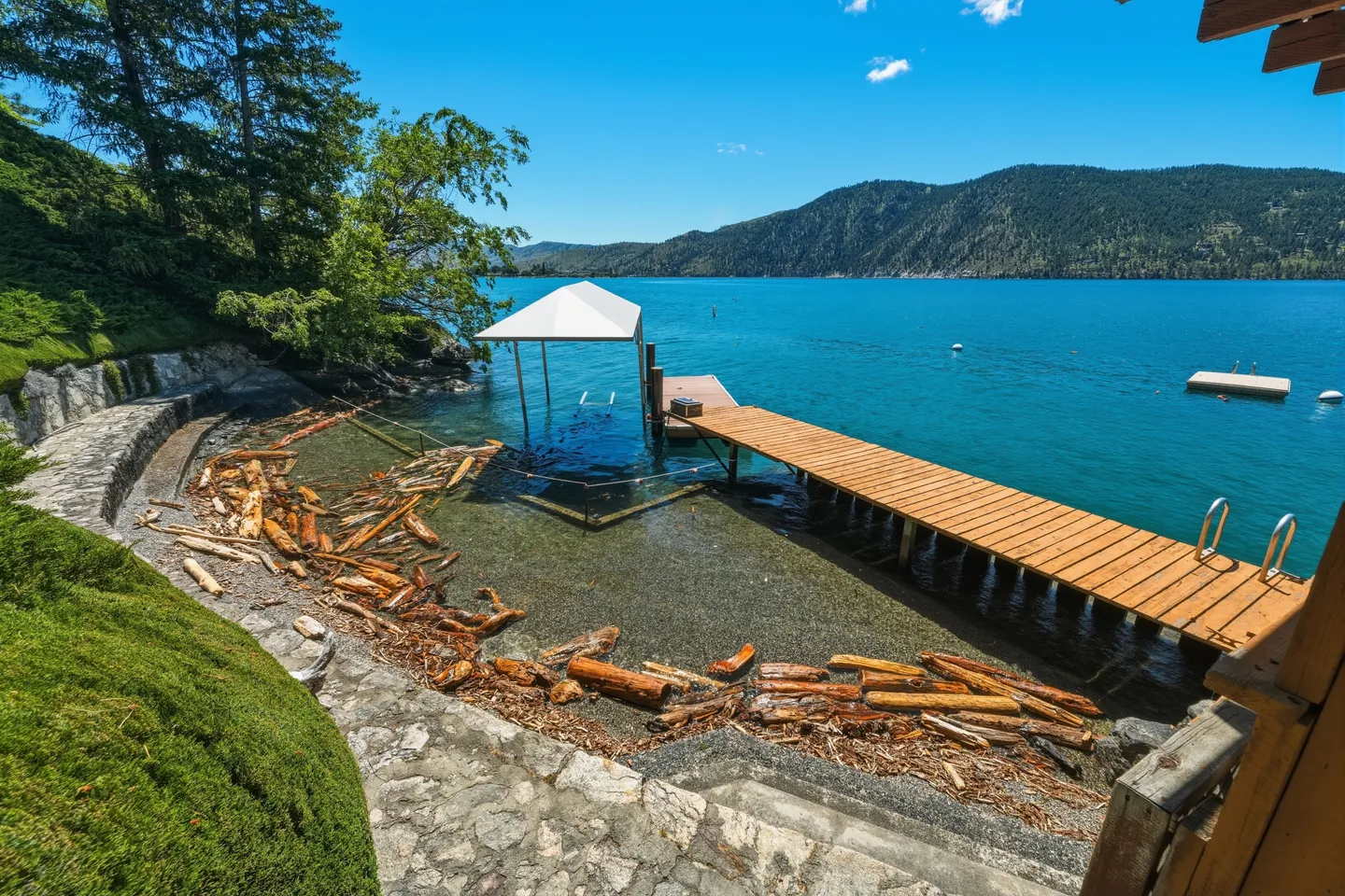 A wooden pier and covered boat lift extend into clear blue water along a curved shoreline with driftwood and mountains in the background.