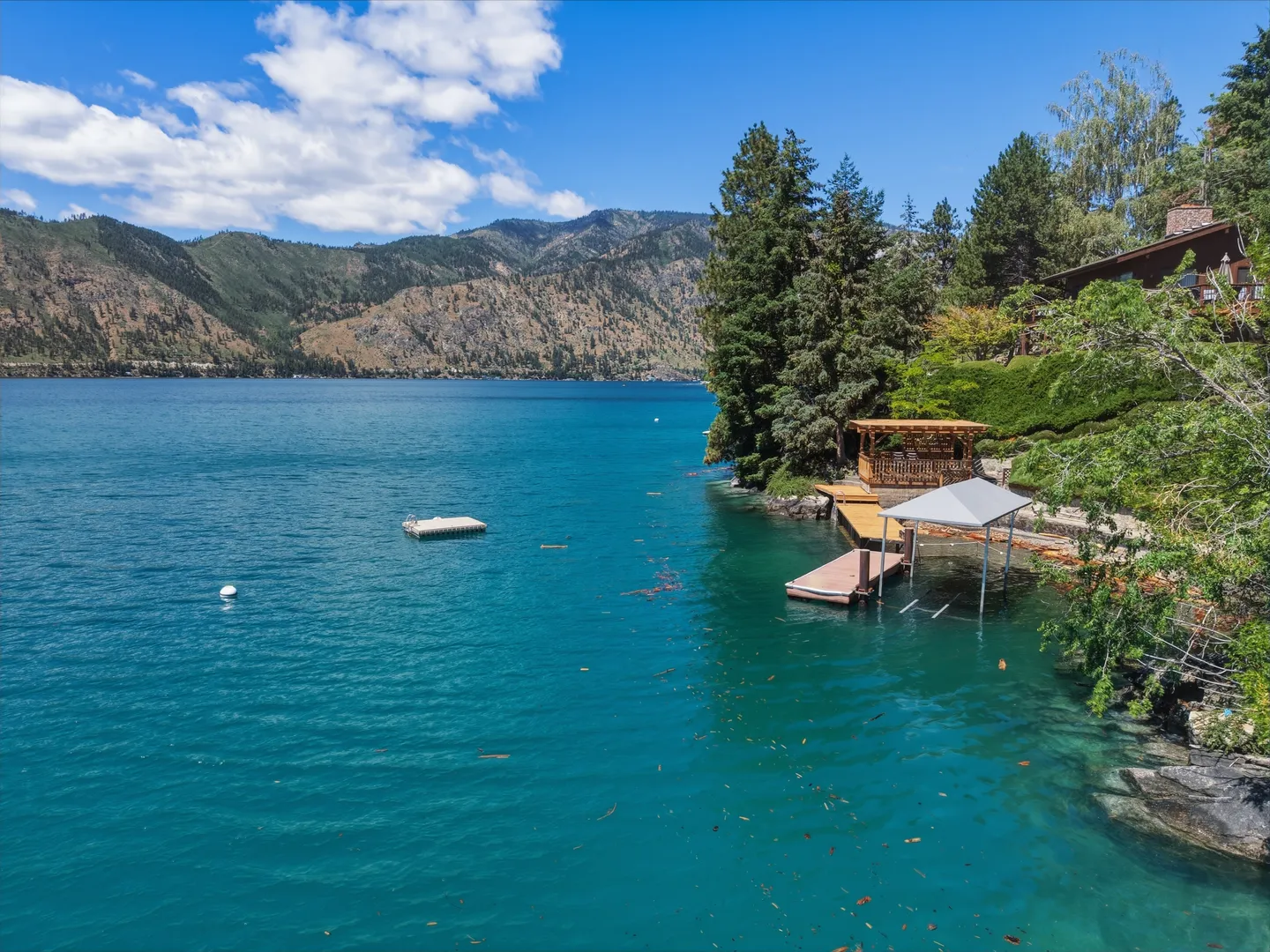 Elevated view of a lakefront shoreline with a private dock, boat lift, gazebo, and swim platform against a mountain backdrop.