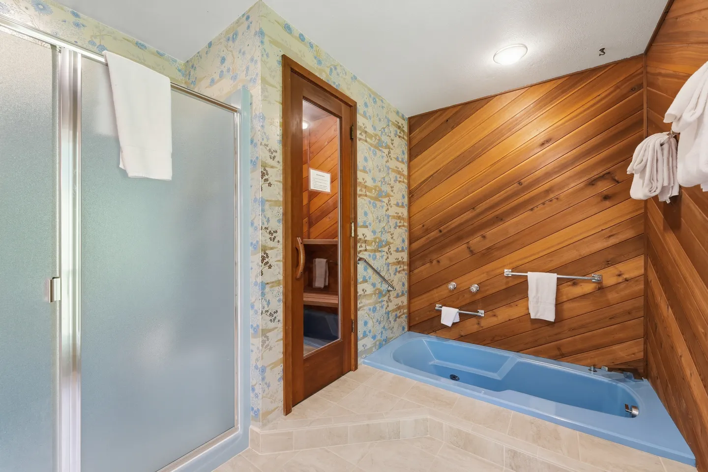 Bathroom featuring a blue sunken tub against diagonal wood paneling, an enclosed sauna with a glass door, and a frosted glass shower stall.