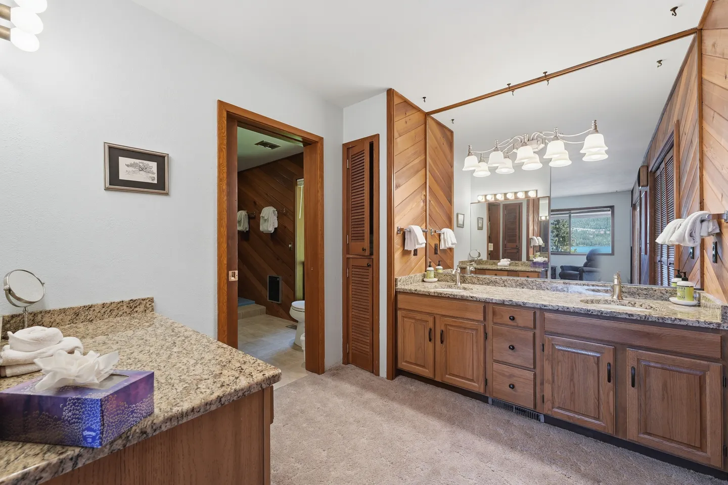 Bathroom vanity area with double sinks, speckled countertops, wood cabinets, a large mirror, and an adjoining toilet room.