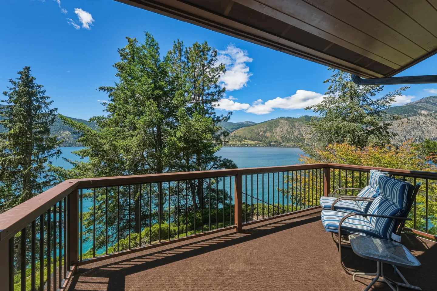 Elevated outdoor balcony with railing and outdoor chairs overlooking a blue lake, mountains, and pine trees.