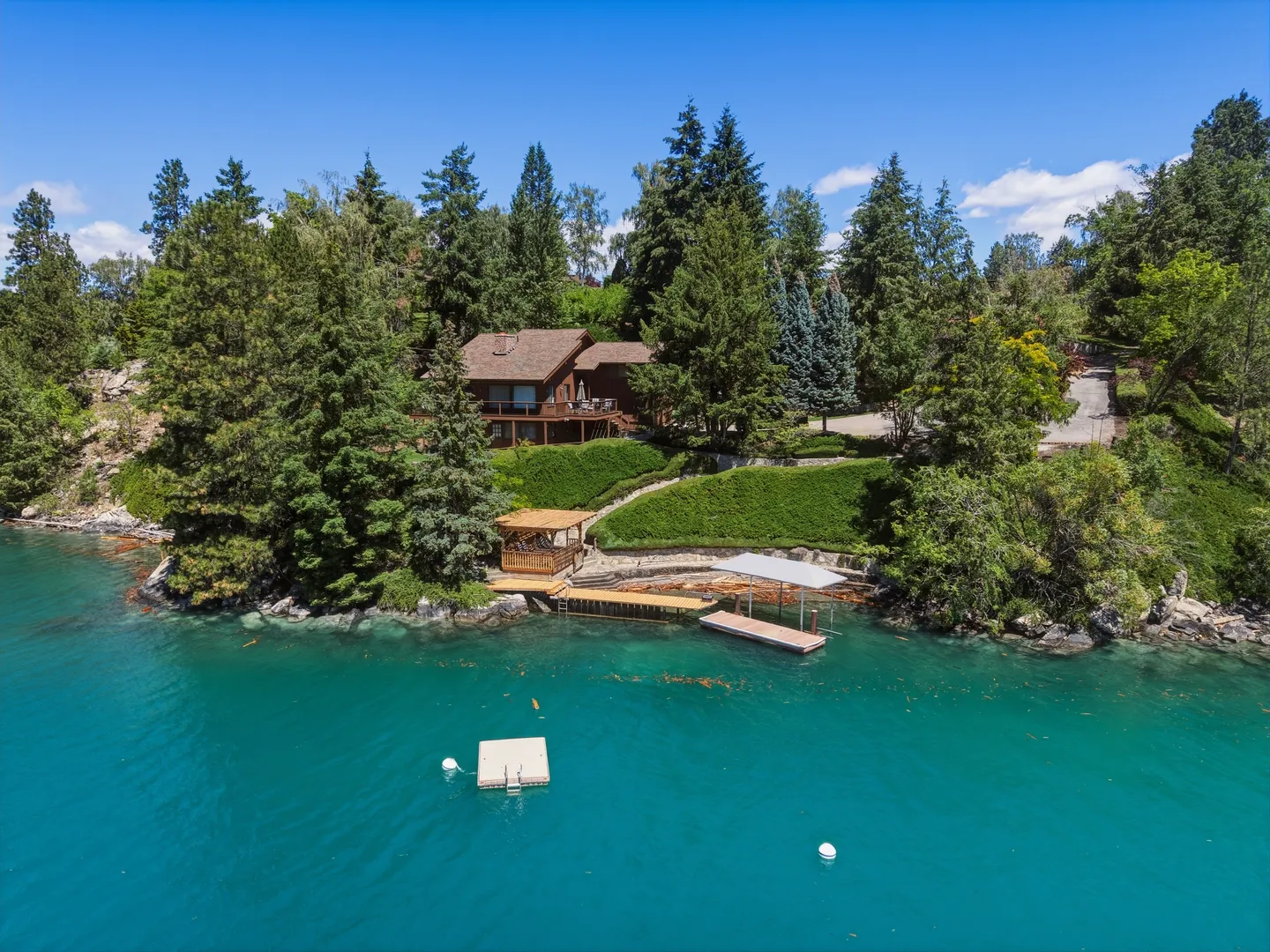 Aerial view of a lakefront house surrounded by tall pine trees with a private dock and swim platform on turquoise water.