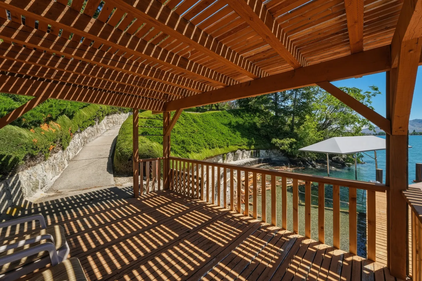 A covered wooden deck or pergola overlooking a lake, with slatted shade overhead and views of green hillside landscaping.